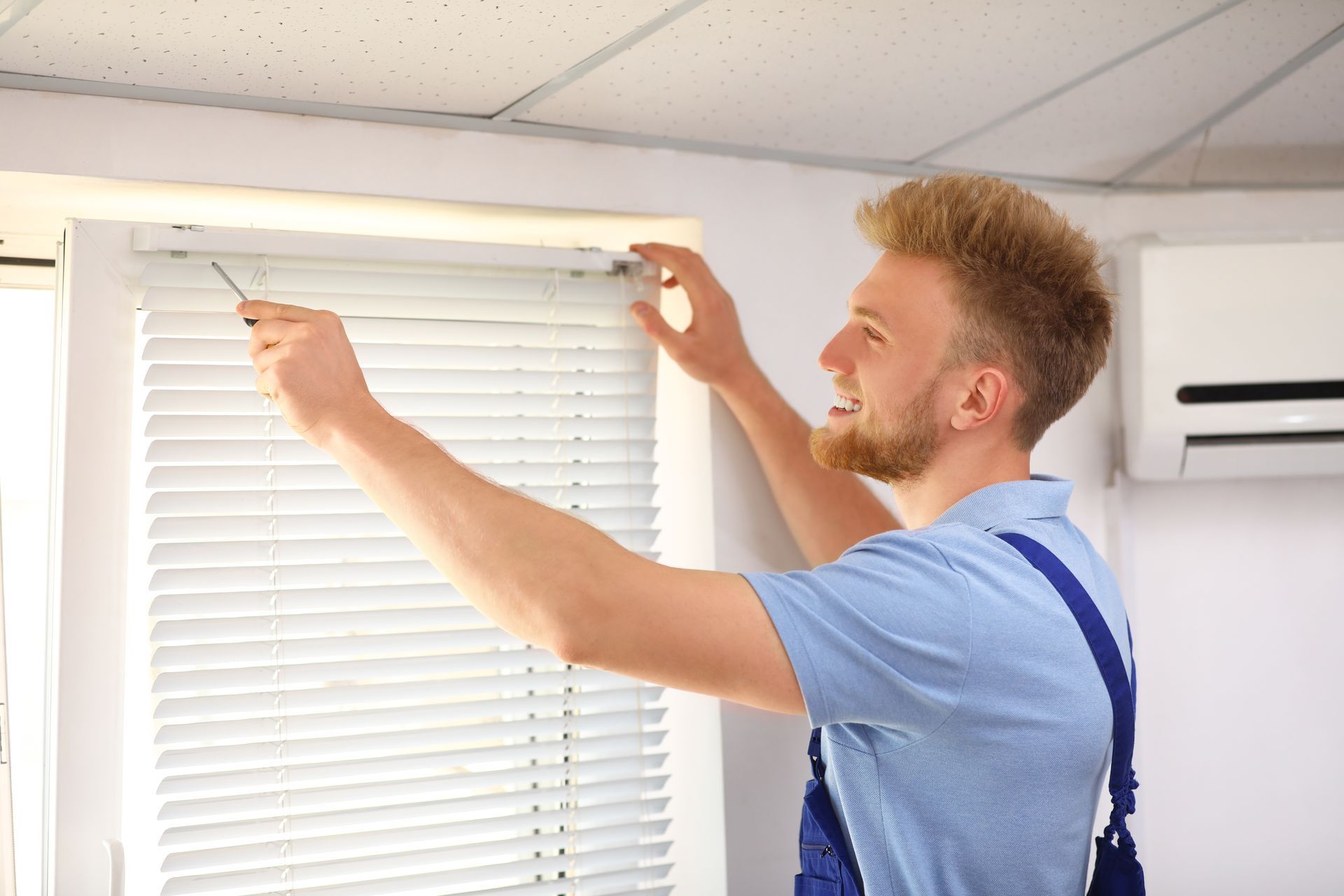 Man adjusting white window blinds in a bright room using a tool and a ladder
