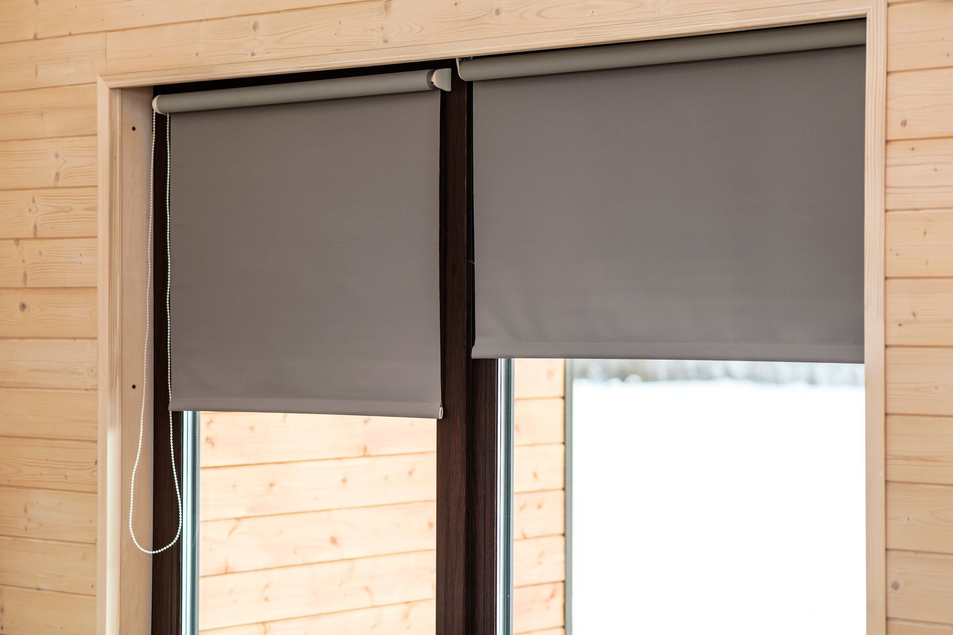Gray roller blinds partially closed over a window in a wooden interior.