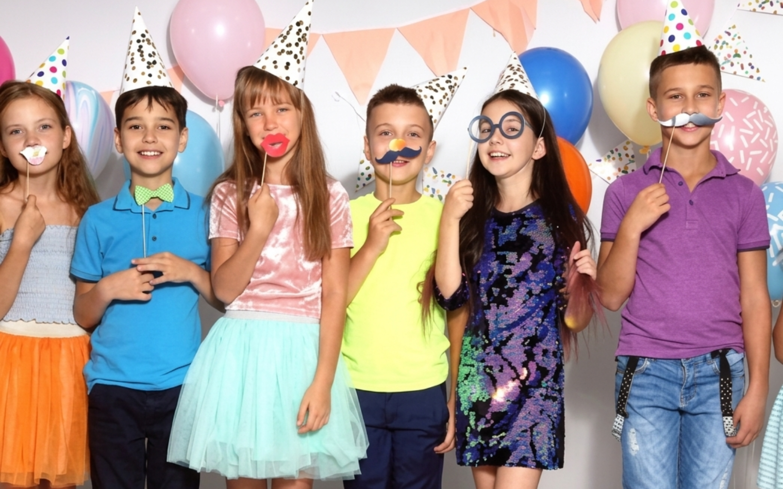 Children wearing party hats and masks stand together holding balloons at a birthday party.