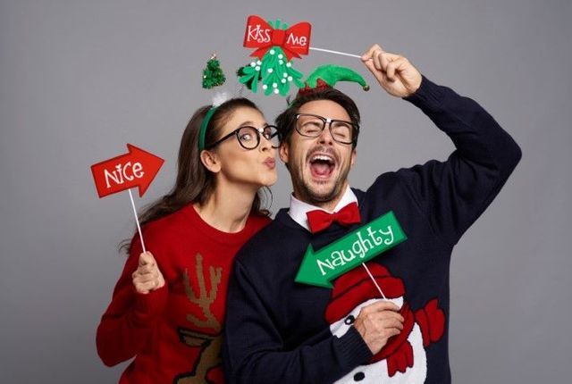 Two people in festive sweaters hold holiday signs and laugh against a gray background.
