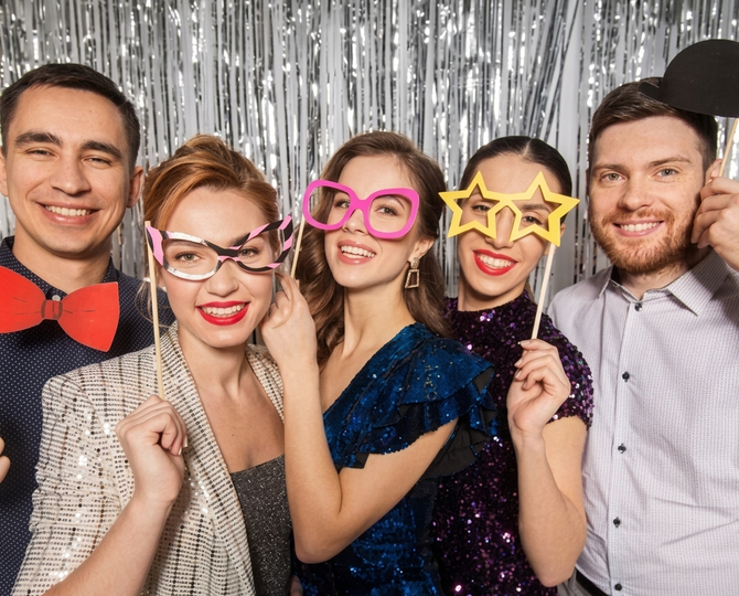 Five friends smiling in festive glasses and bow tie props in front of silver tinsel curtains