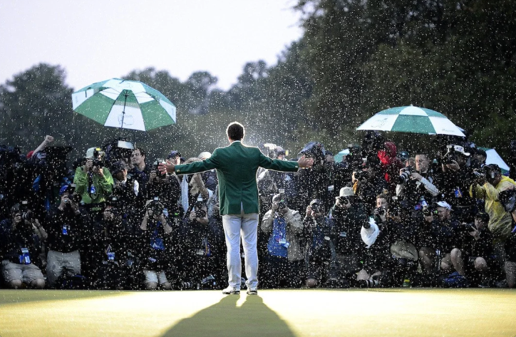 A man in a green jacket is standing in front of a crowd of people holding umbrellas.