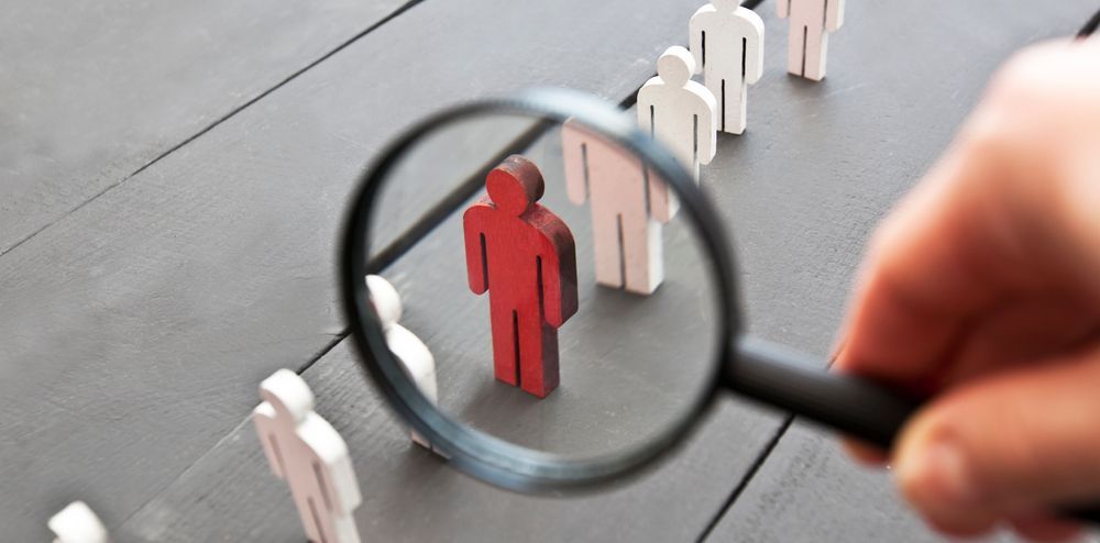 a person holding a magnifying glass over a red toy man in a line with white toy men