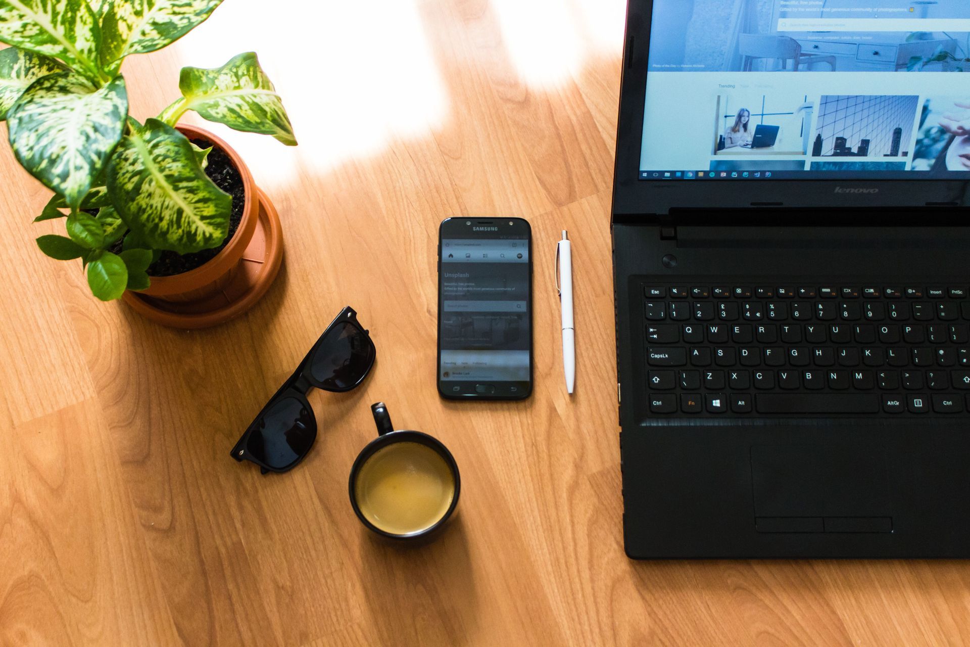 Top-down desk setup with laptop, smartphone, coffee, sunglasses, pen, and potted plant on a wooden table