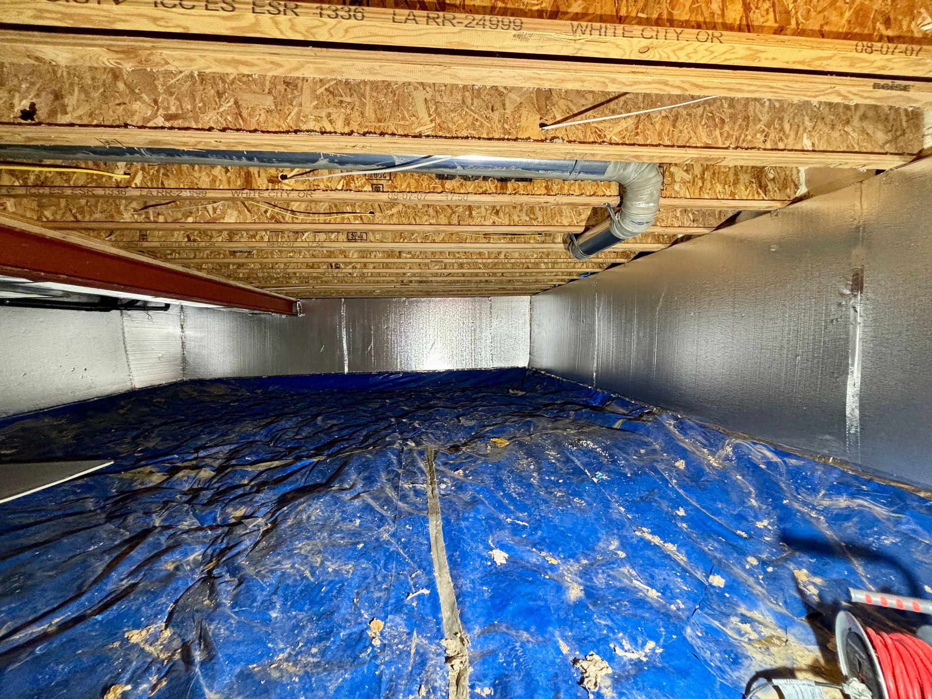 Crawlspace with blue tarp, silver insulation, exposed beams, and a pipe.