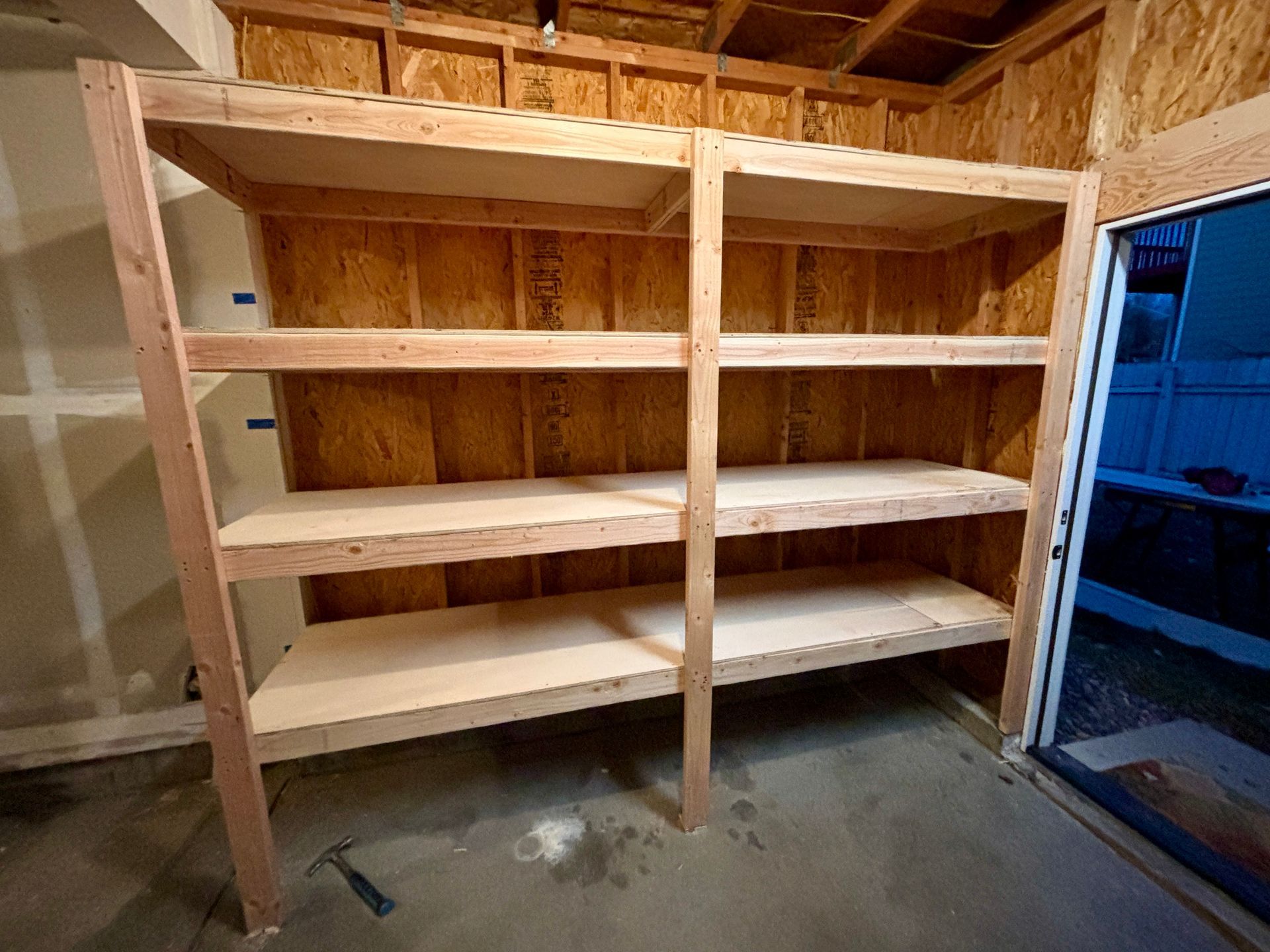 Wooden shelving unit in a garage, against plywood walls and concrete floor.
