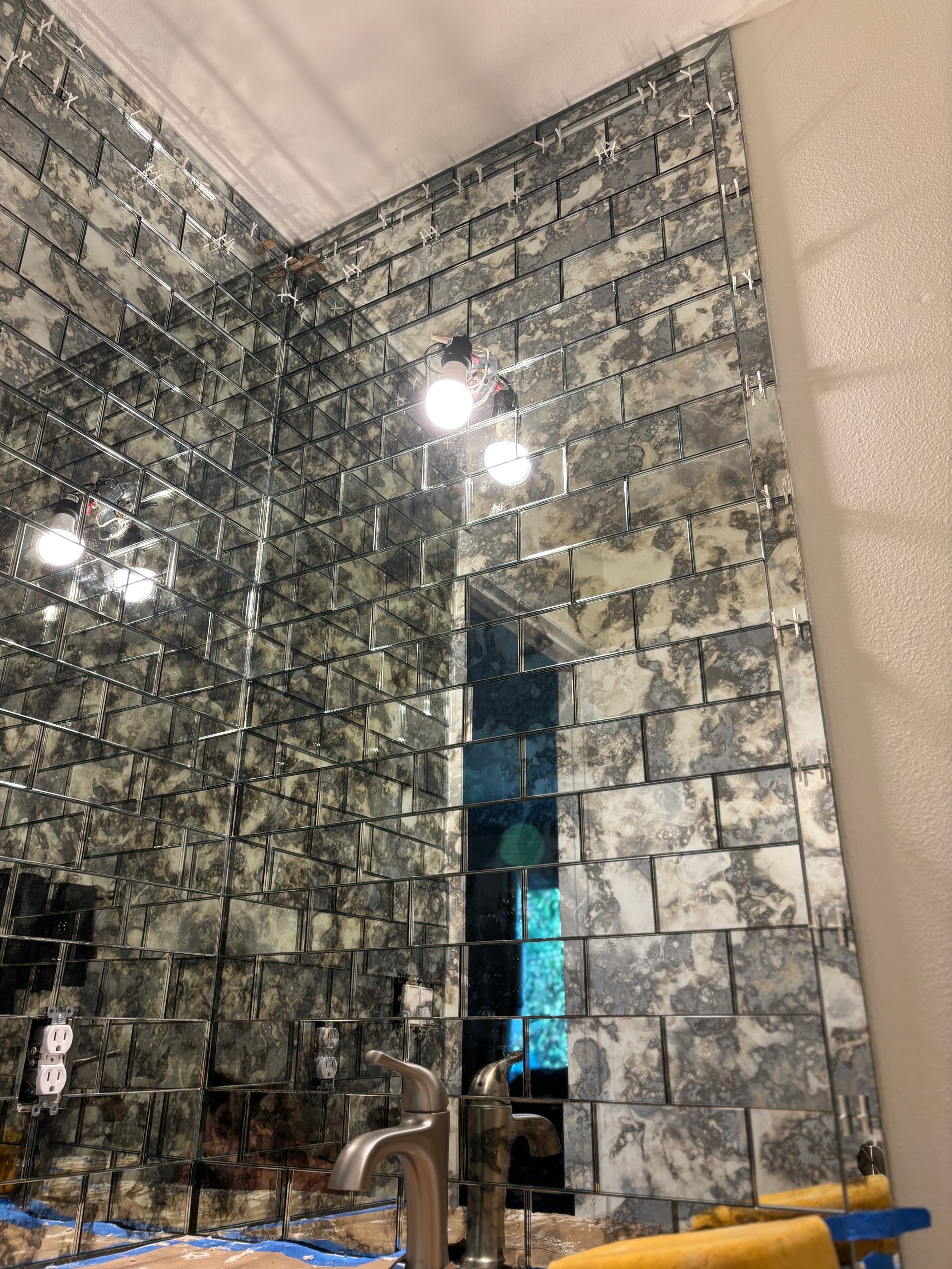 Mirror-tiled walls in a bathroom with a silver faucet, reflecting the room and light fixtures above.