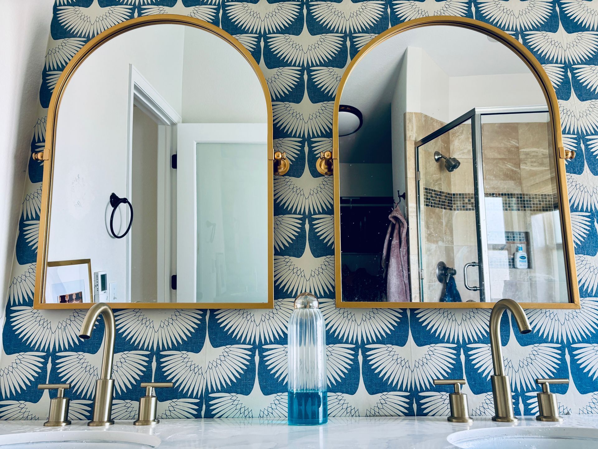 Bathroom with two arched gold mirrors, blue patterned wallpaper, and gold faucets.