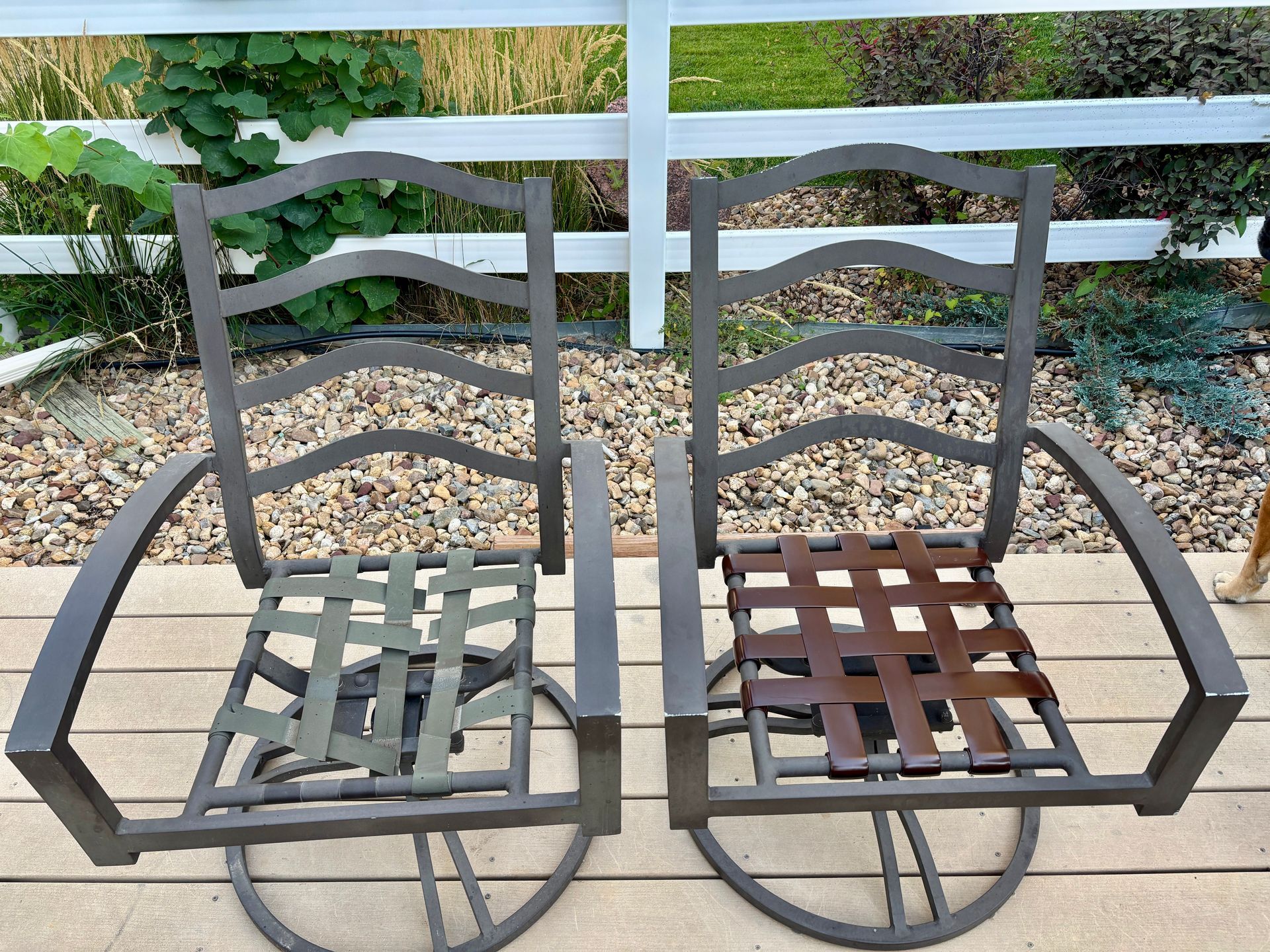 Two metal outdoor chairs, one with green straps, the other with brown, on a deck with a white fence backdrop.
