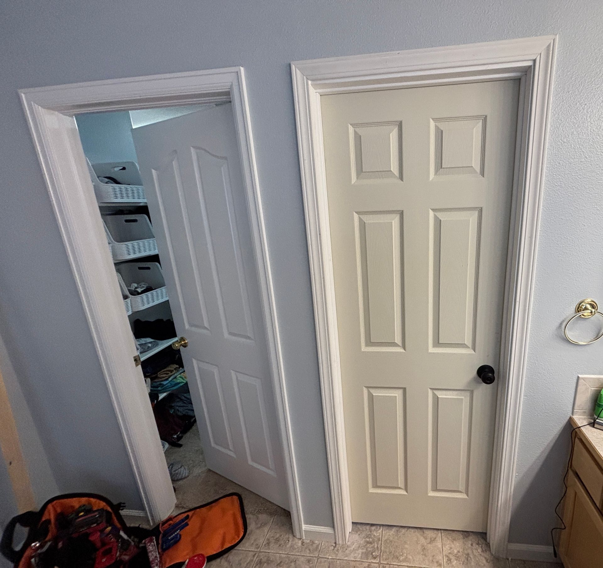 Two white doors; one open revealing shelves, the other closed. Light beige walls, tile flooring.