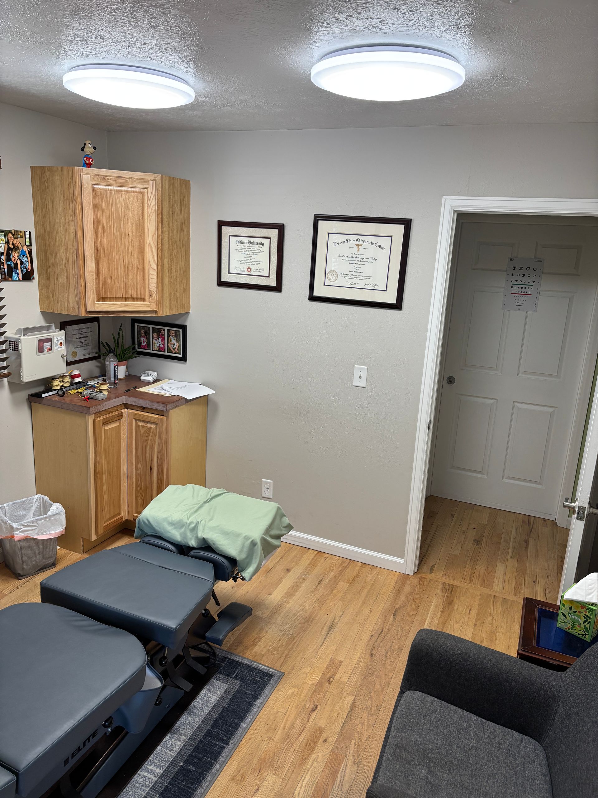 Chiropractor's office interior: treatment table, cabinets, diplomas, door, and grey walls.