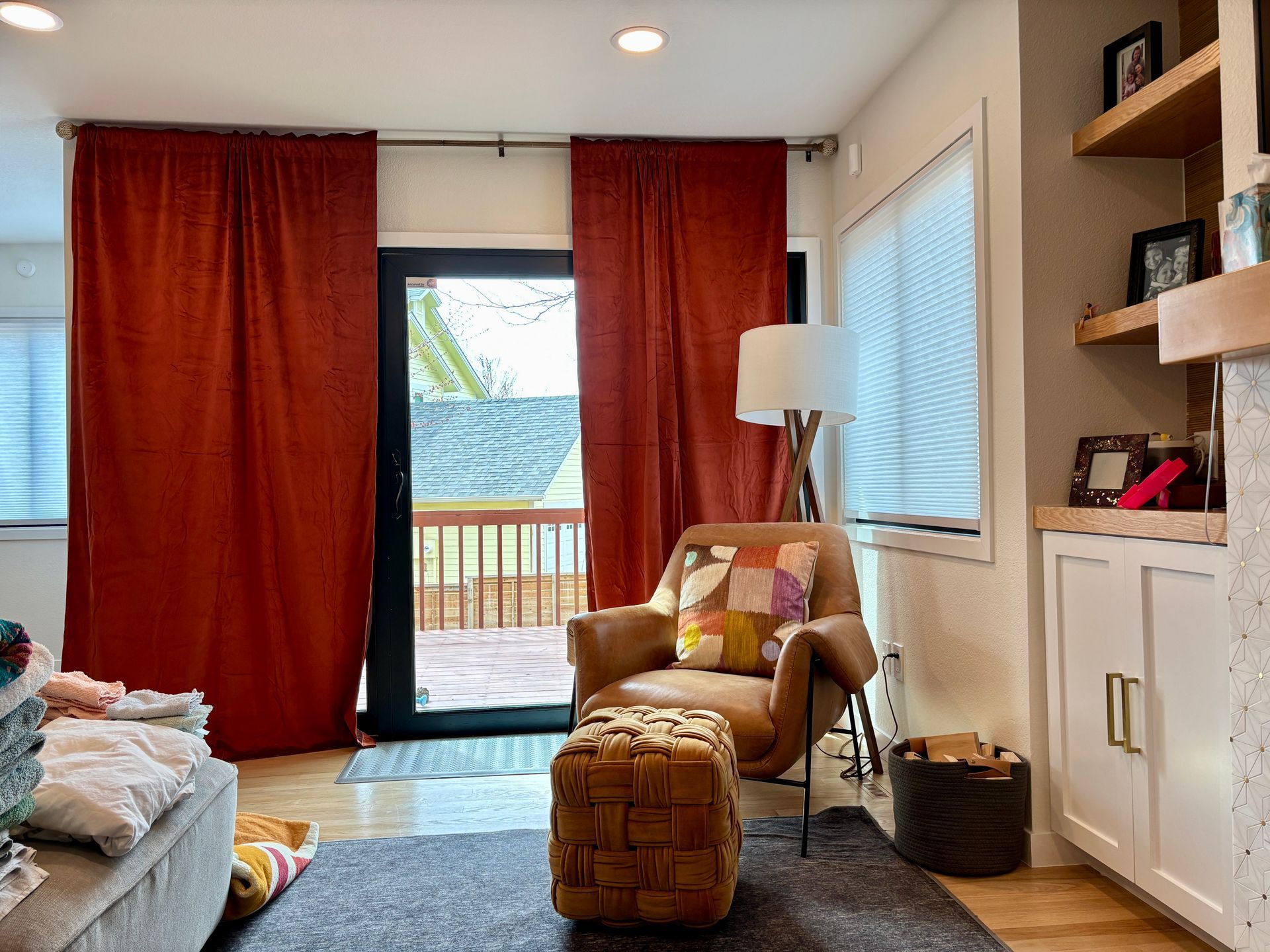 Cozy living room with rust-colored curtains framing a door to a deck, armchair, and built-in shelves.