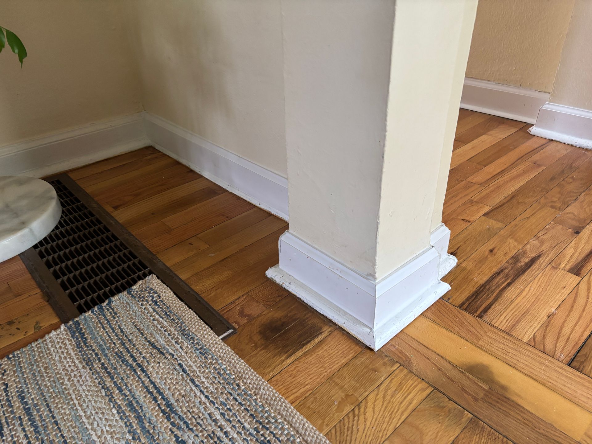 Wooden parquet floor with a floor vent and a square cream-colored pillar. A rug is partially visible.