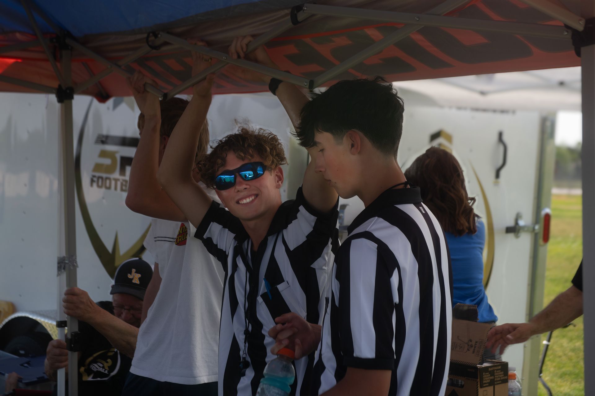 Three people wearing black and white striped official shirts stand under a canopy, with one smiling in blue sunglasses.