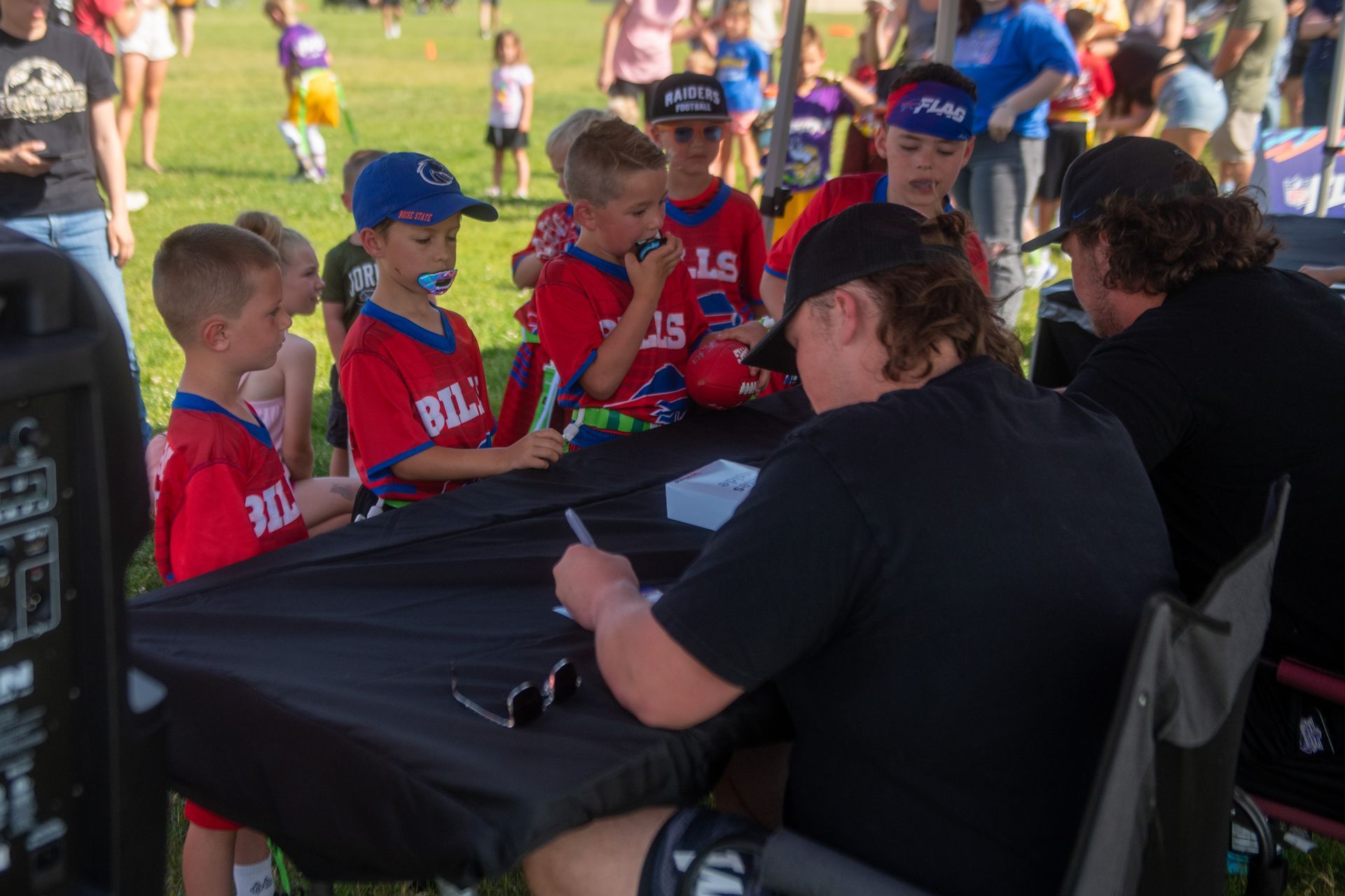 A professional athlete in a black shirt signs autographs for children wearing red sports jerseys at an outdoor event.
