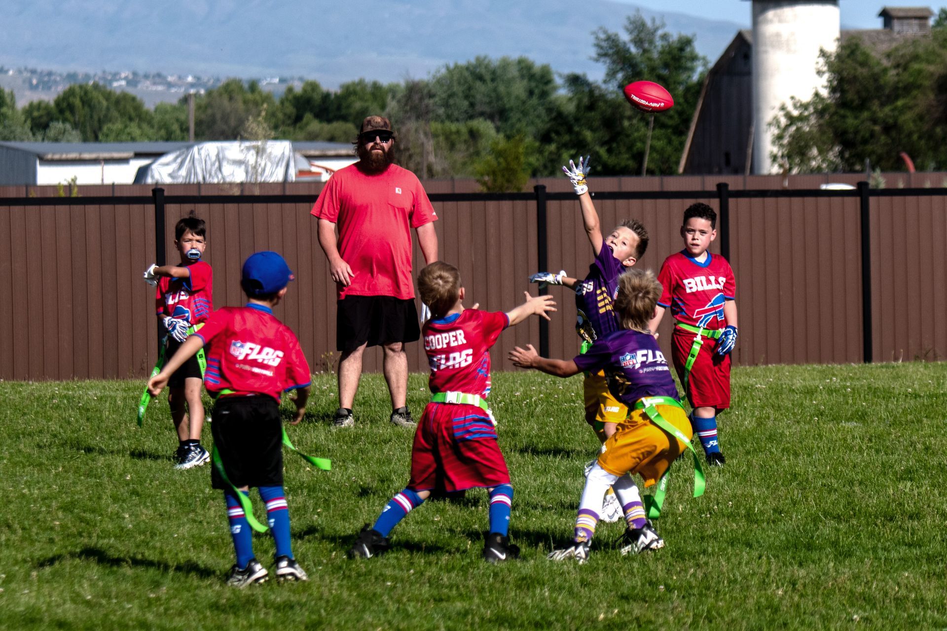 A group of children in sports jerseys playing flag football on a grassy field, with a coach watching in the background.