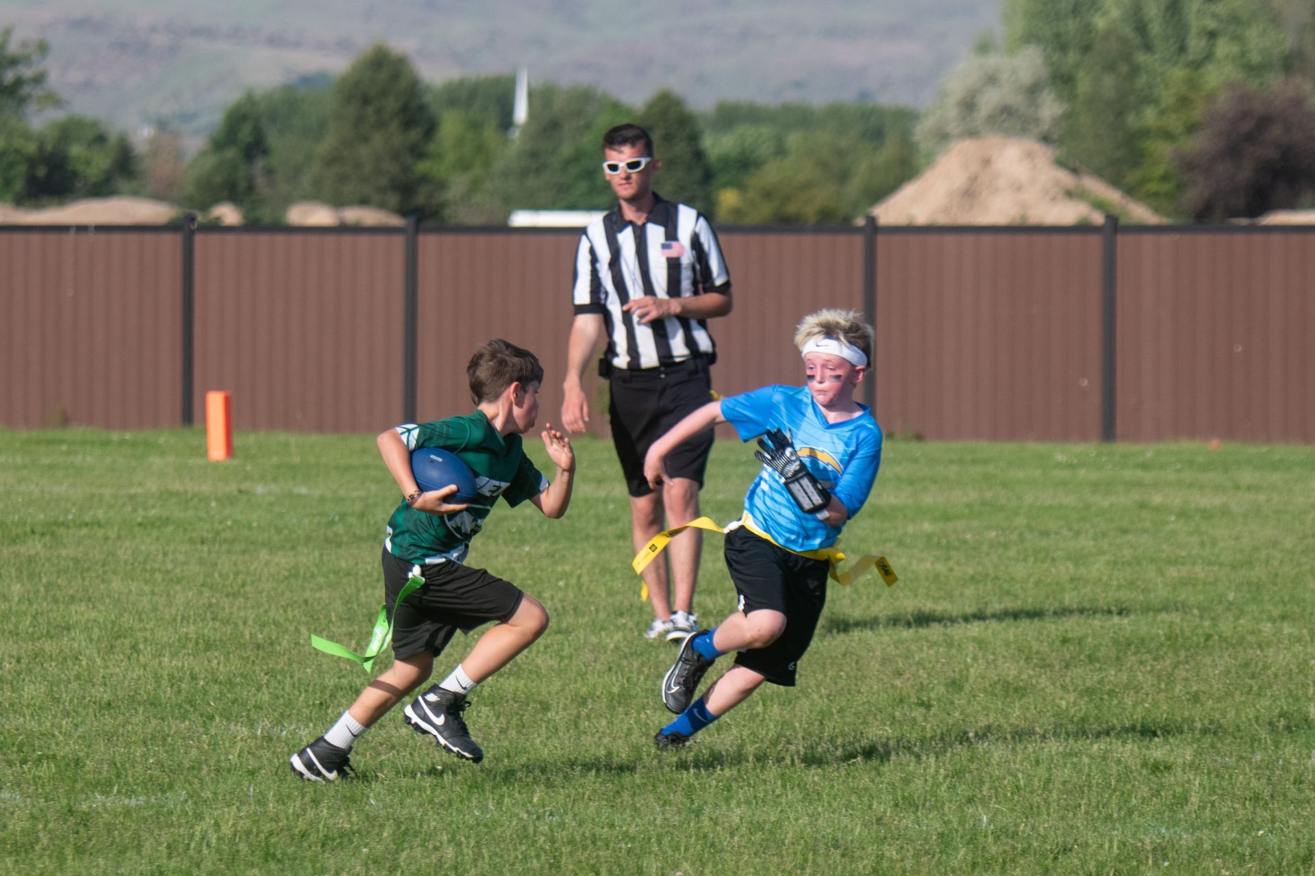Two children play flag football on a grassy field while a referee in a striped shirt observes the play.
