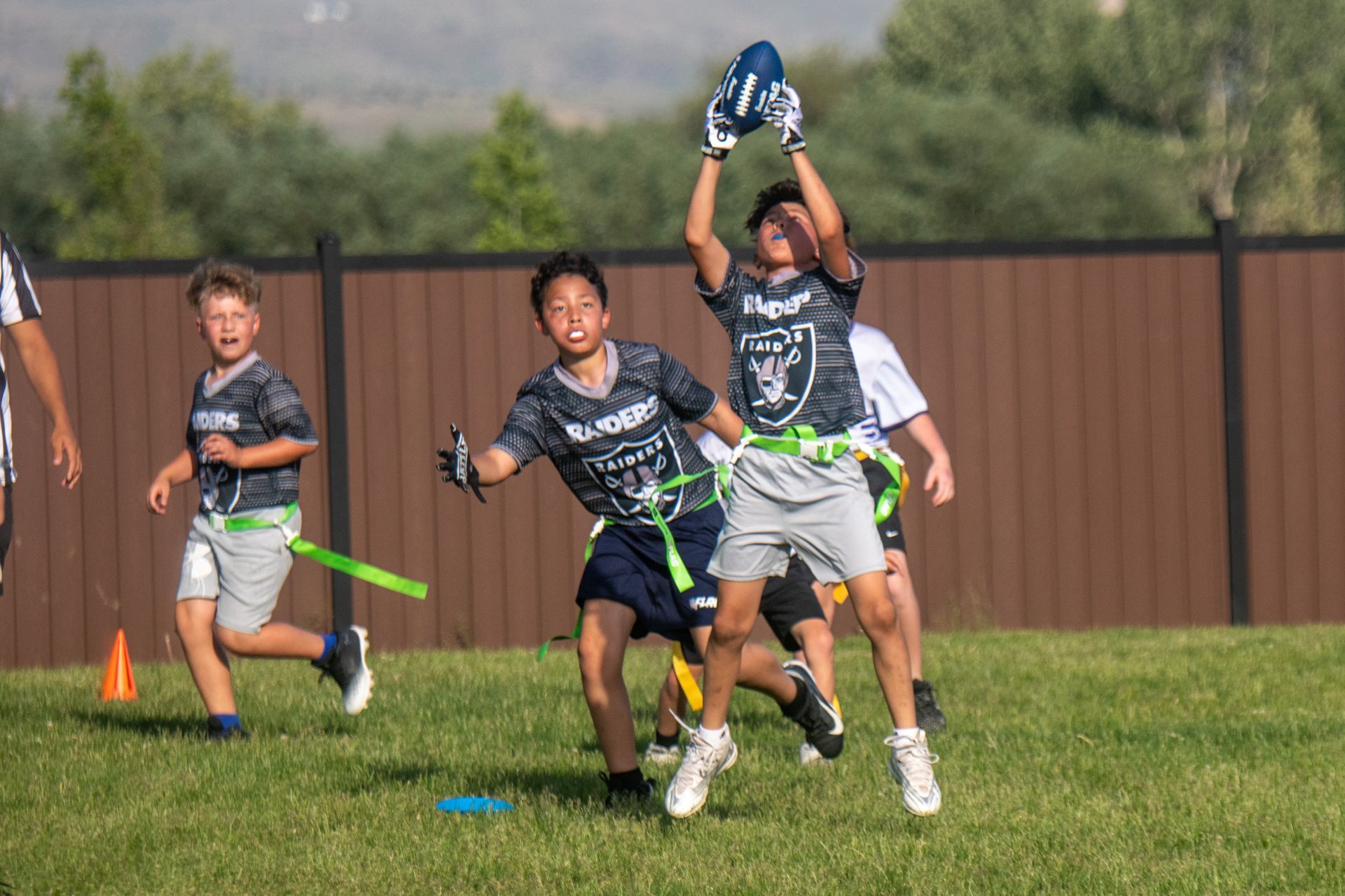 Children play a flag football game on a grassy field, with one player jumping to catch a blue ball.