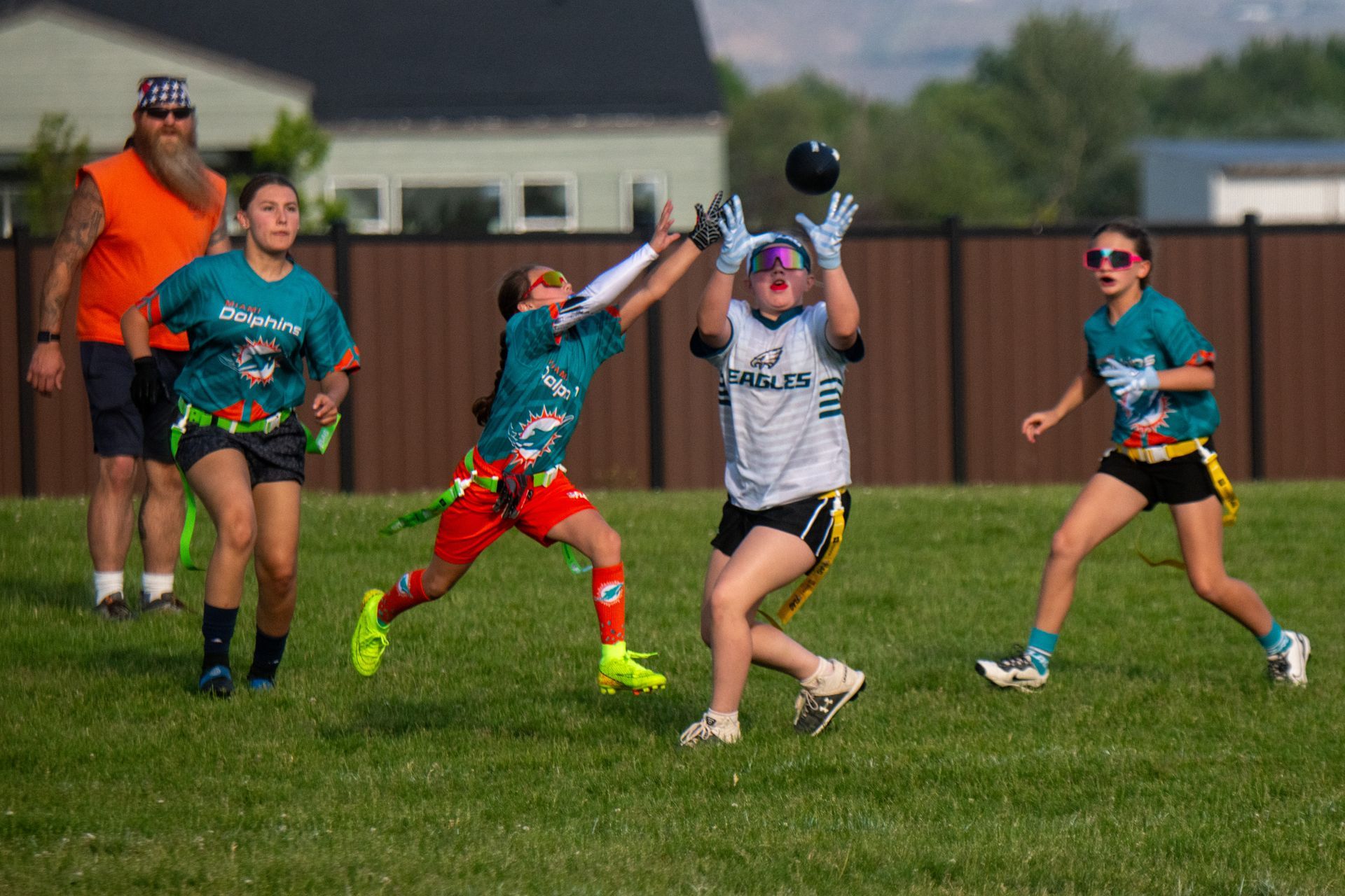 Four players in jerseys compete for a black football on a grassy field with an adult observer in the background.