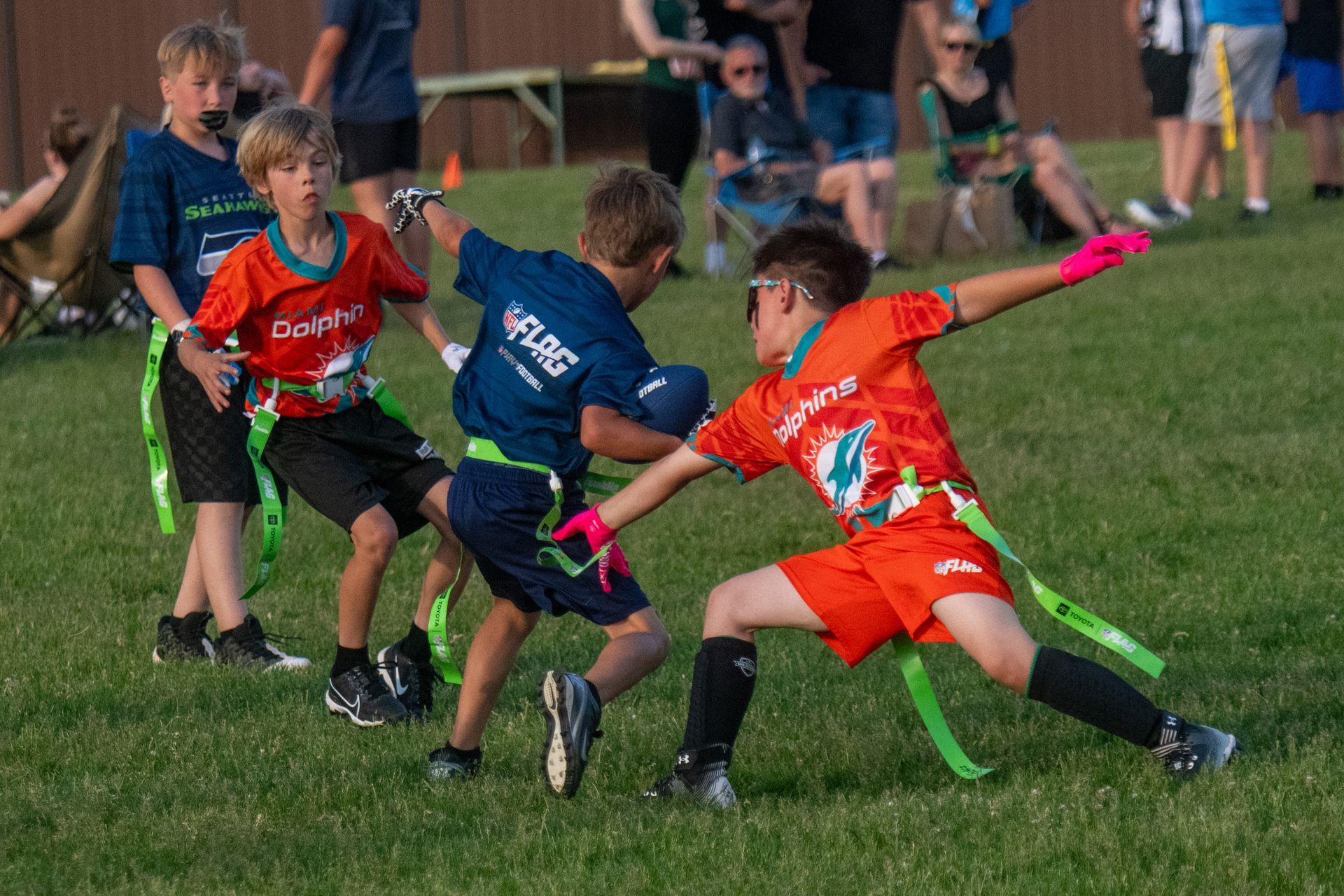 Three children in colorful sports uniforms play flag football on a grassy field.