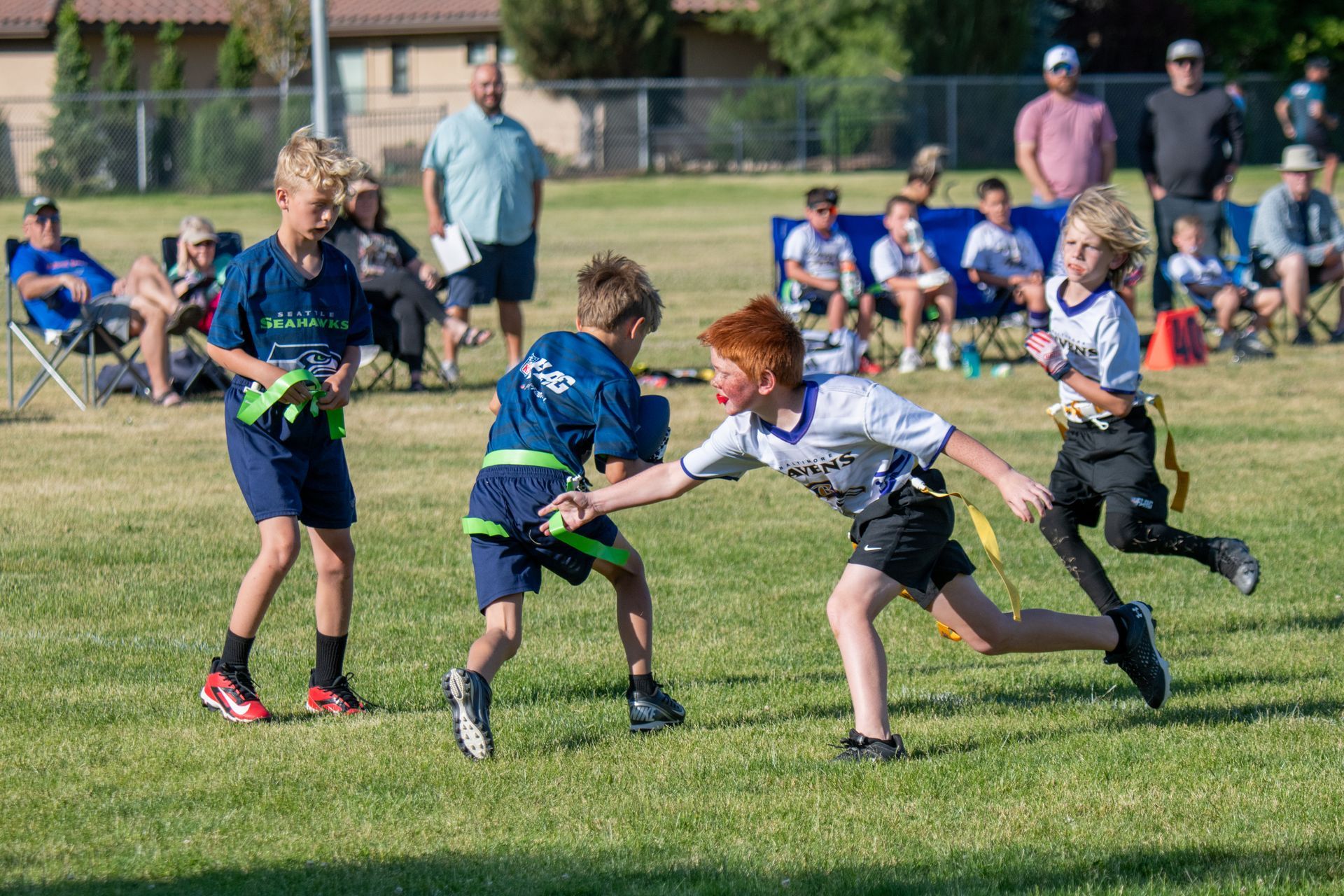 A flag football game in a park with players in blue and white jerseys running and reaching for flags on a grass field.