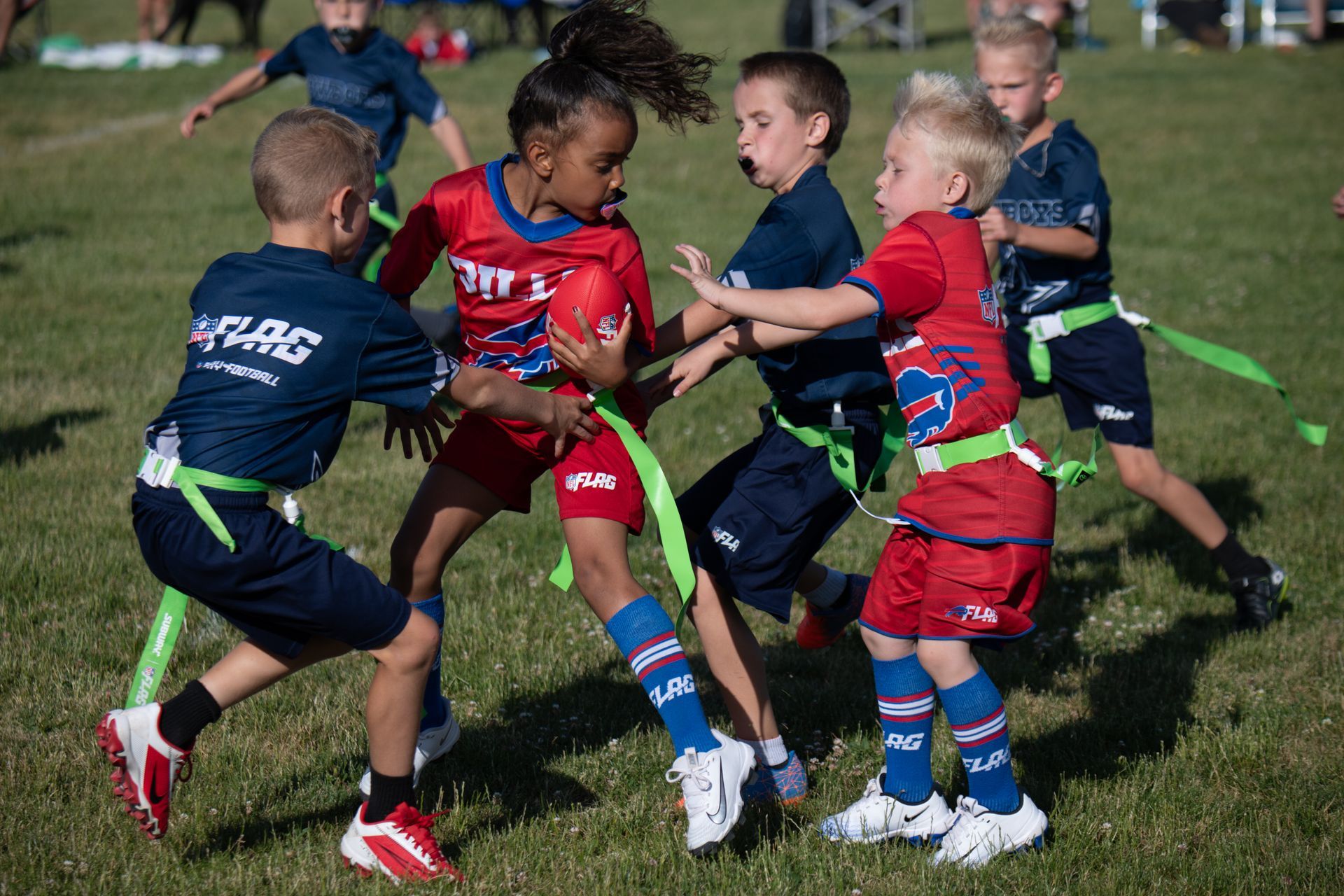 Children in red and navy uniforms play flag football on a grassy field, focusing on a player running with the ball.