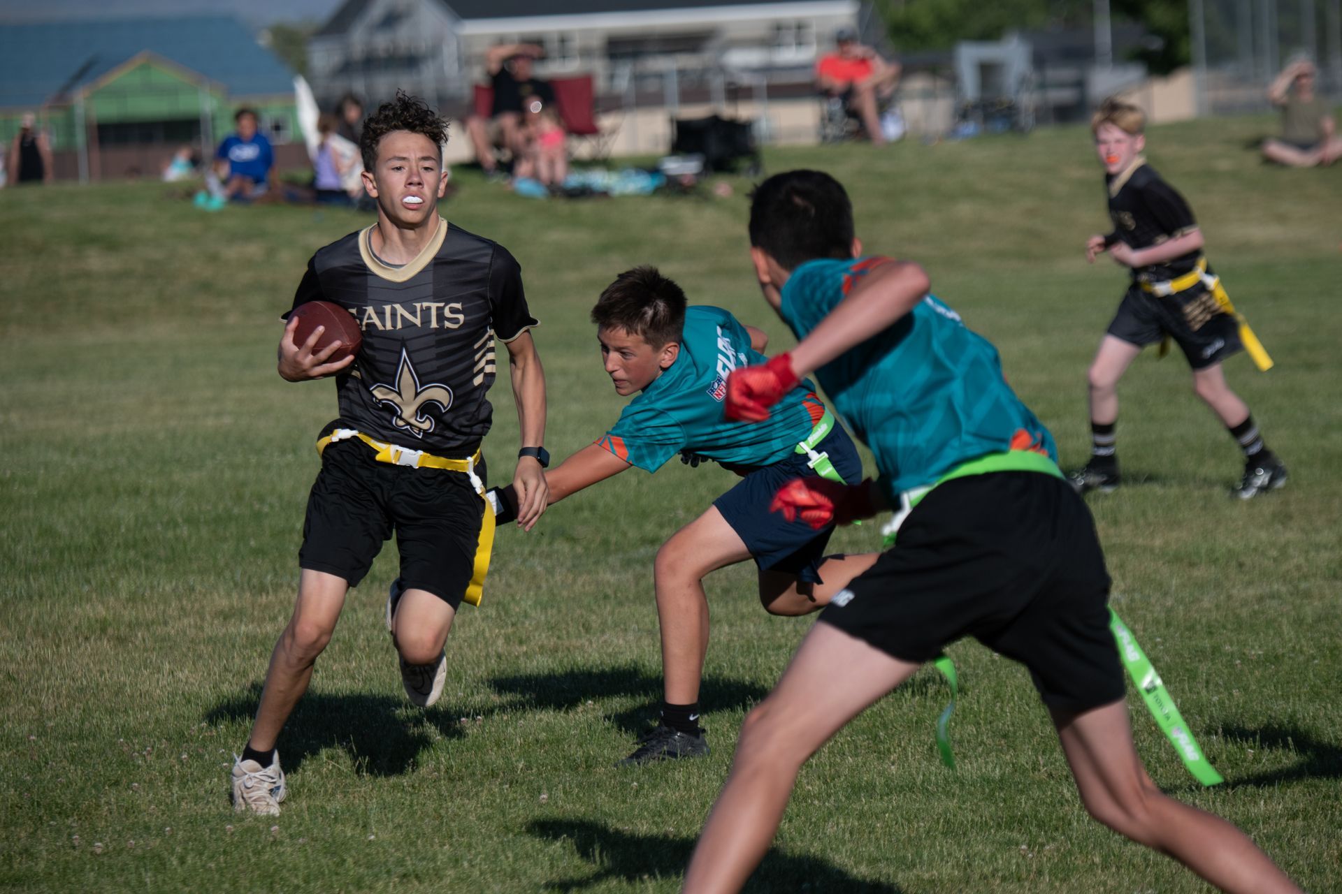 A player in a Saints jersey runs with a football while being pursued by two opponents in teal shirts on a grass field.