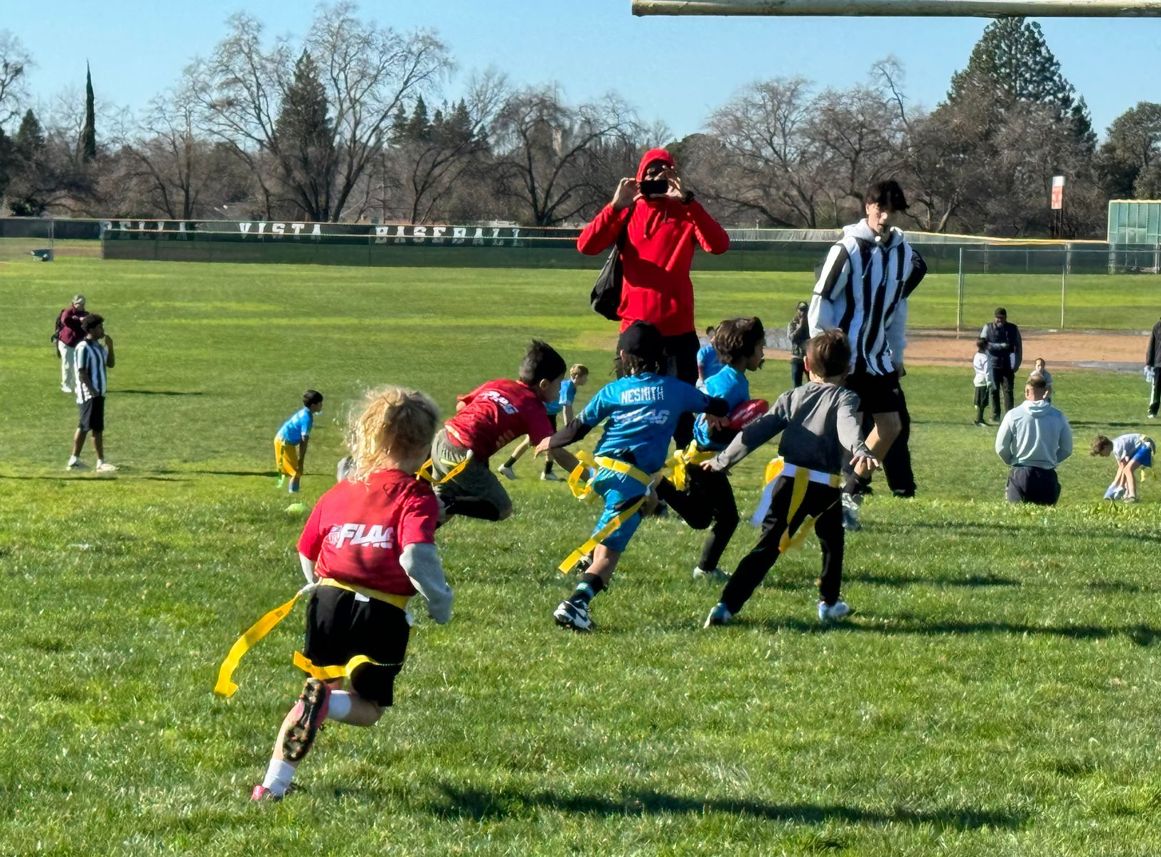 Children in red and blue jerseys playing a game of flag football on a sunny grass field with coaches nearby.