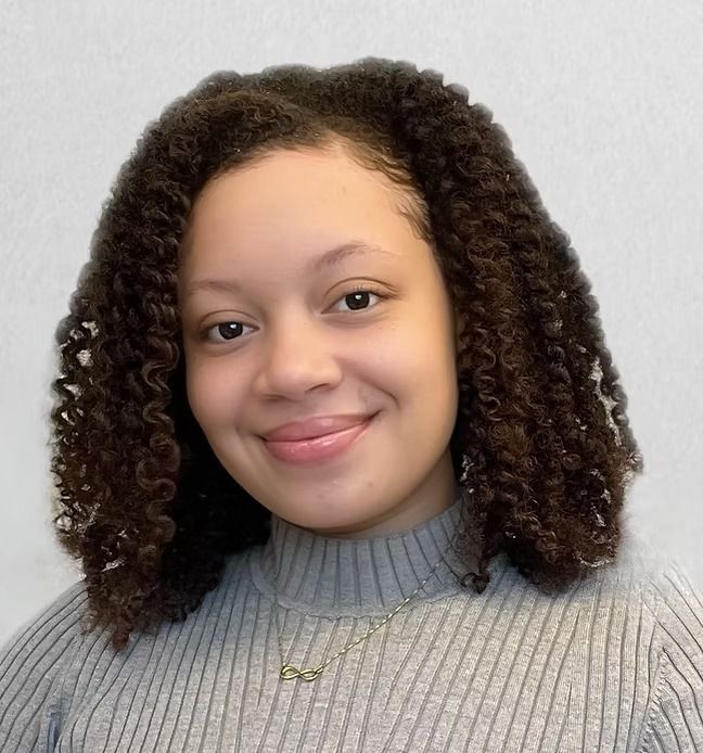 A woman with curly hair is wearing a grey sweater and a necklace.