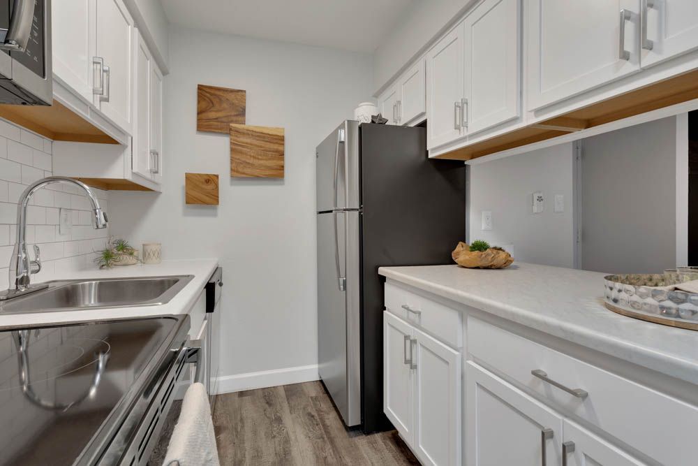 Narrow white galley kitchen with stainless fridge, sink, white cabinets, and wood-look floor