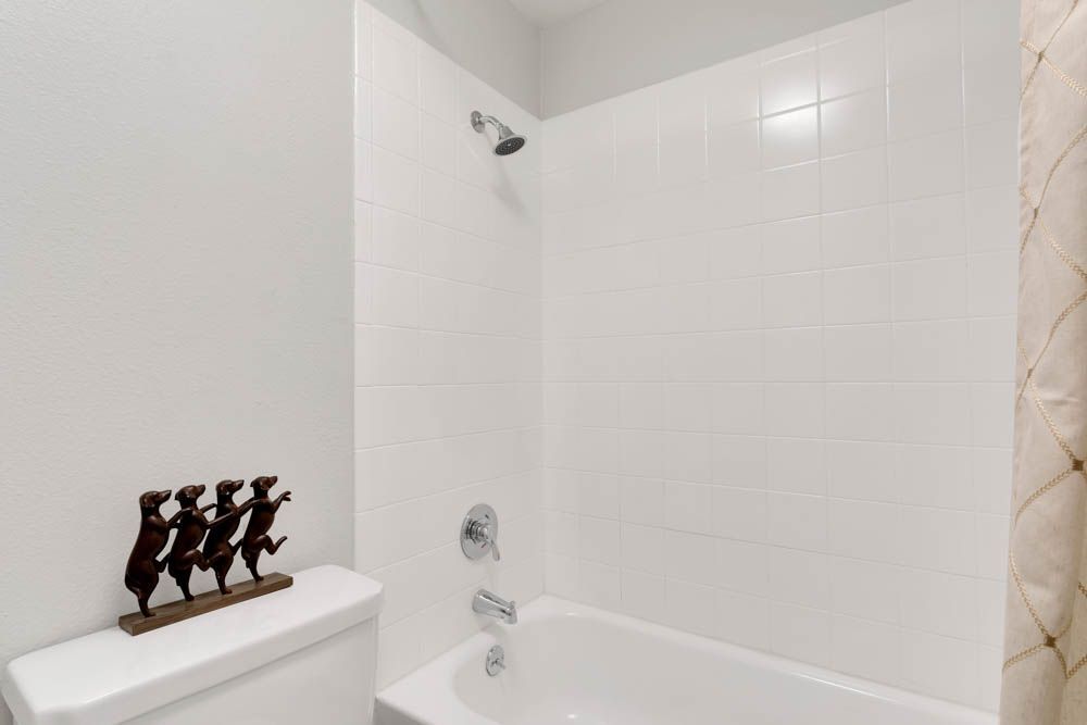 White bathroom with a tiled bathtub shower, wall-mounted spout, and dark decorative figures on the toilet tank.