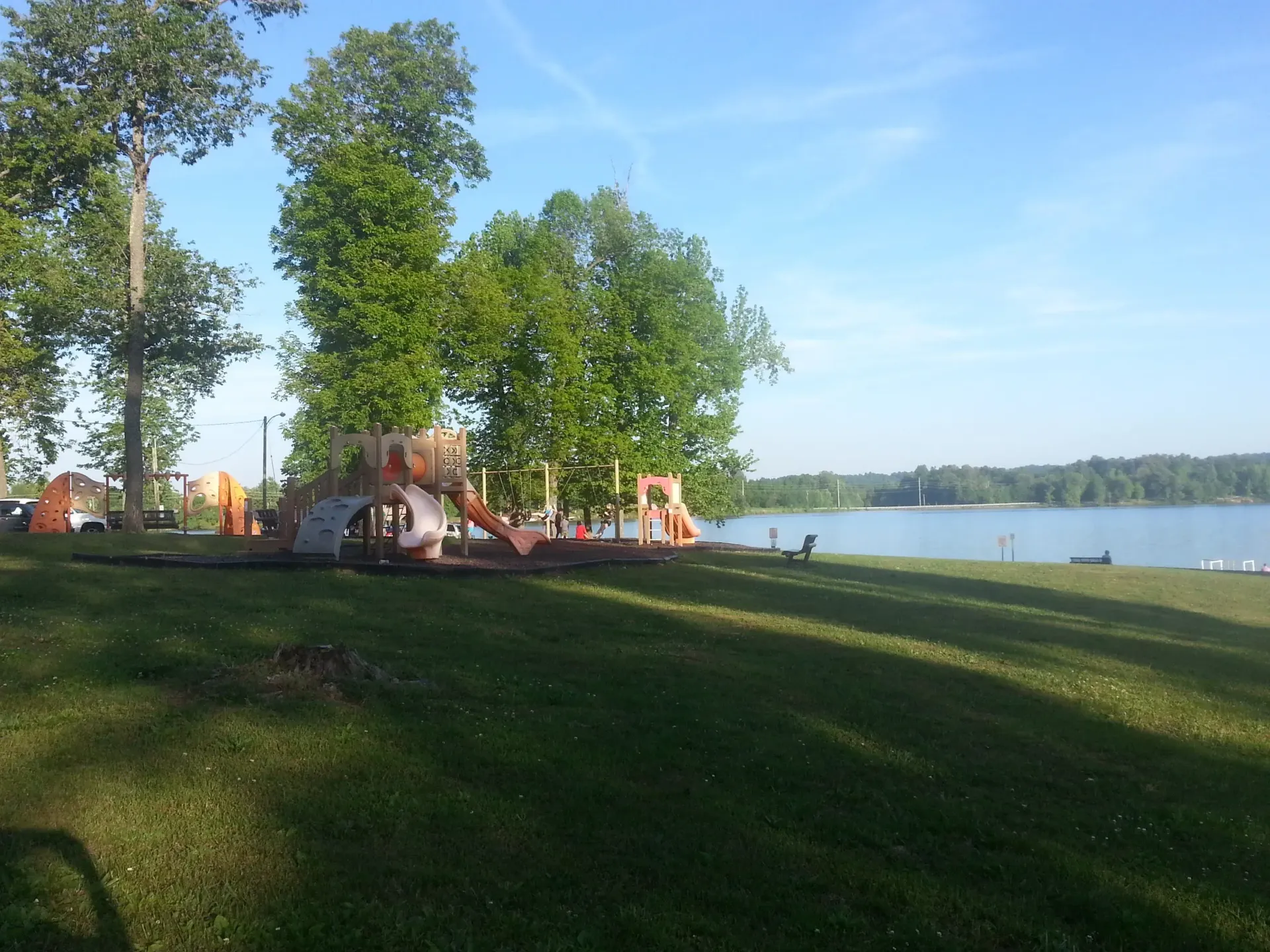Playground with slides and climbing structures on a grassy lakefront in Earlington, Kentucky, with a lake and wooded shoreline in the background