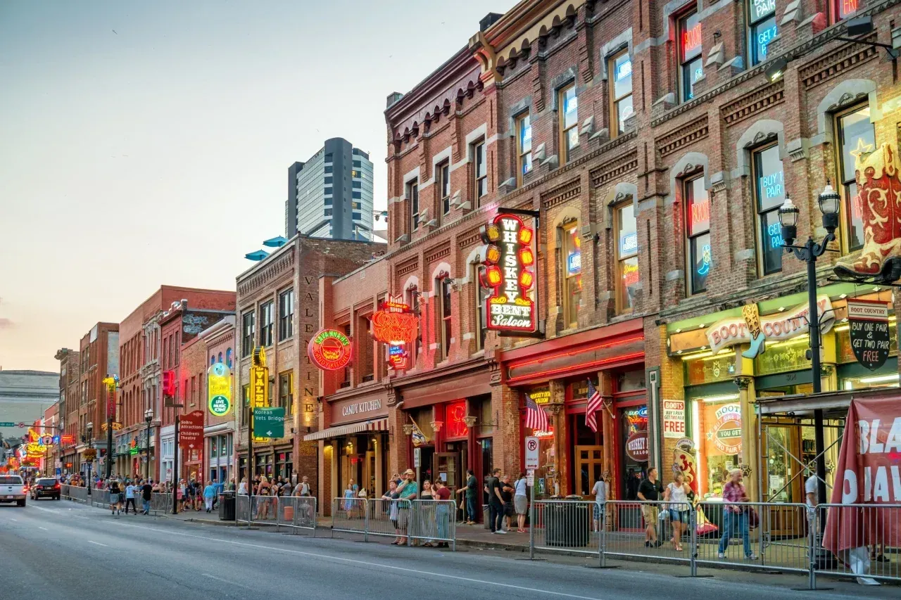 Broadway Street in downtown Nashville, Tennessee at dusk, lined with neon-lit honky-tonk bars including Whiskey Bent Saloon and Boot Country