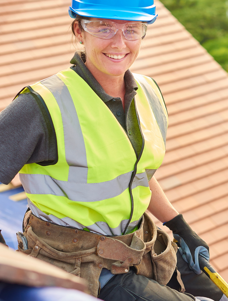 A woman wearing a hard hat and safety vest is sitting on a roof.