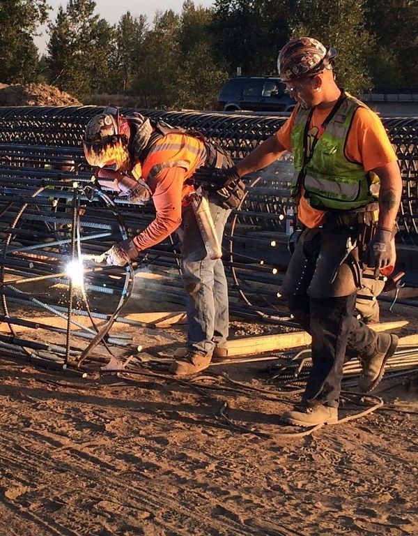 Two construction workers are welding a metal structure on a dirt road.