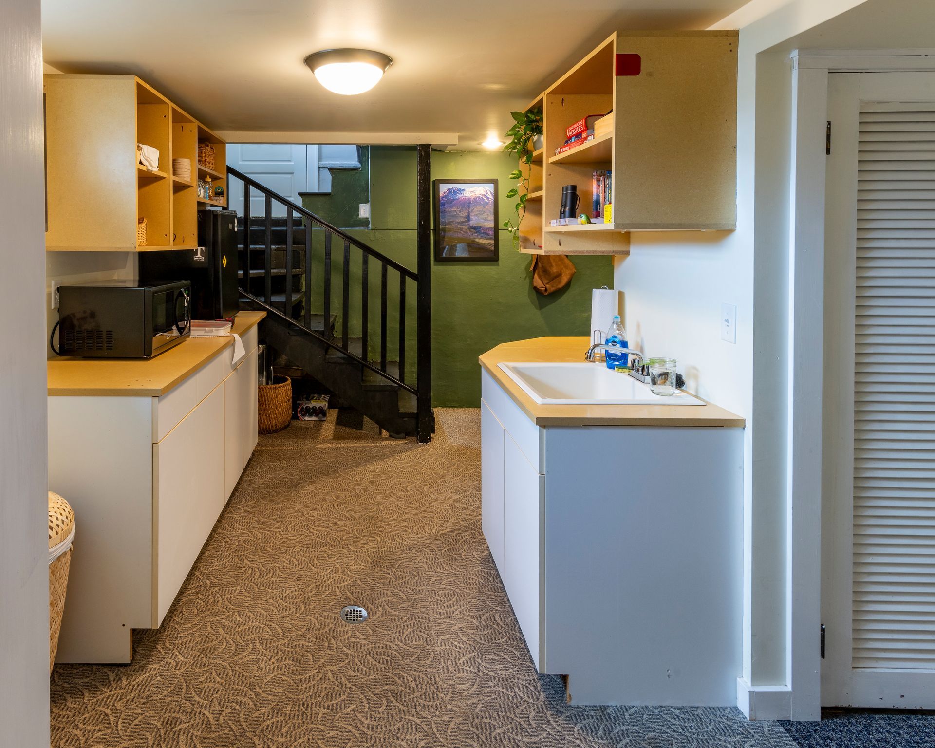 Narrow kitchen hallway with white cabinets, green wall, staircase, and a sink under warm ceiling light