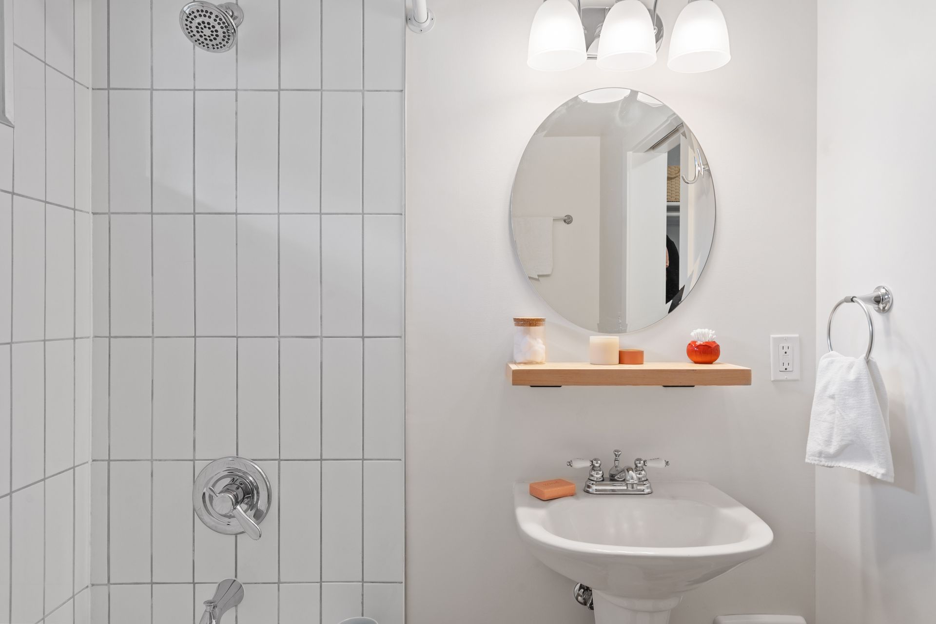 White bathroom with tiled shower, pedestal sink, round mirror, shelf, and towel rack