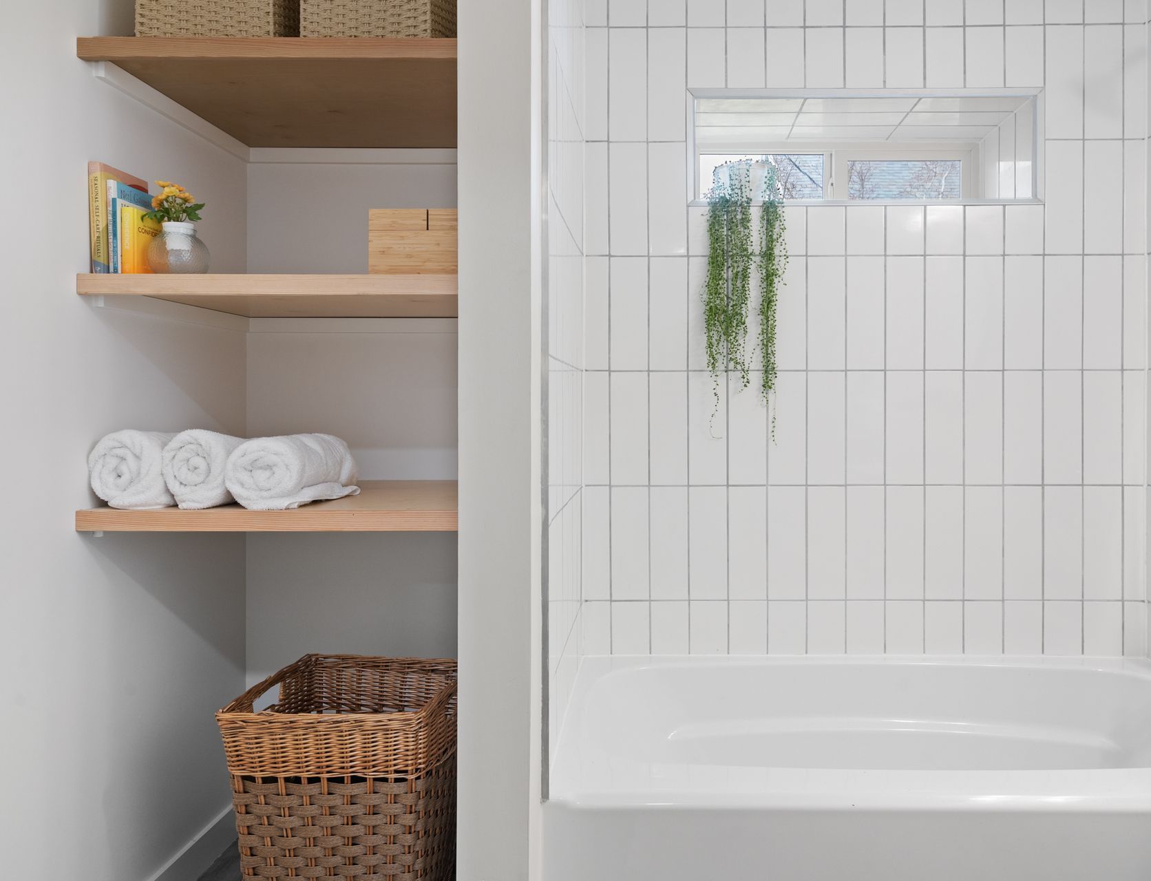 Bathroom shelving beside white tiled shower and bathtub, with baskets, towels, books, and a hanging plant.