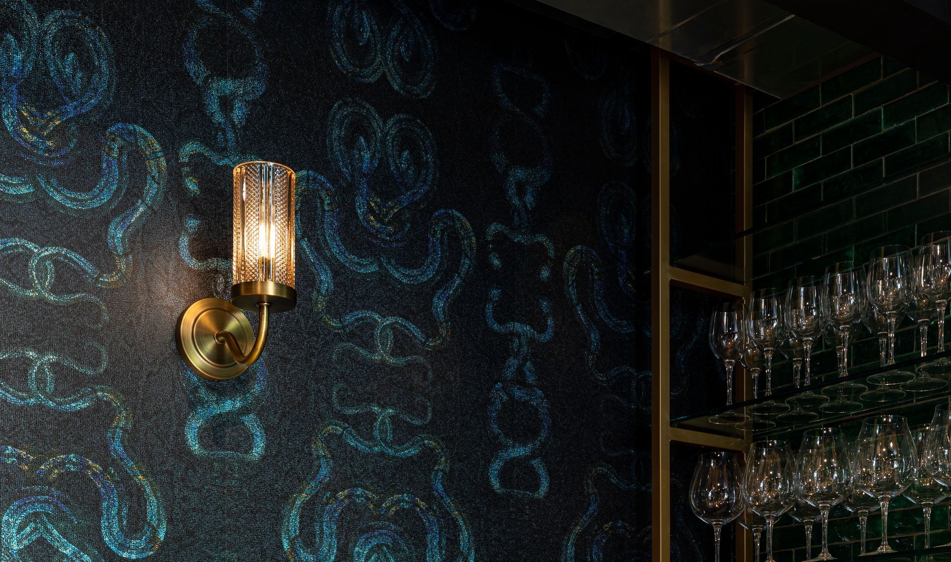 Dimly lit bar wall with blue patterned wallpaper, brass sconce, and glass shelves of stemware