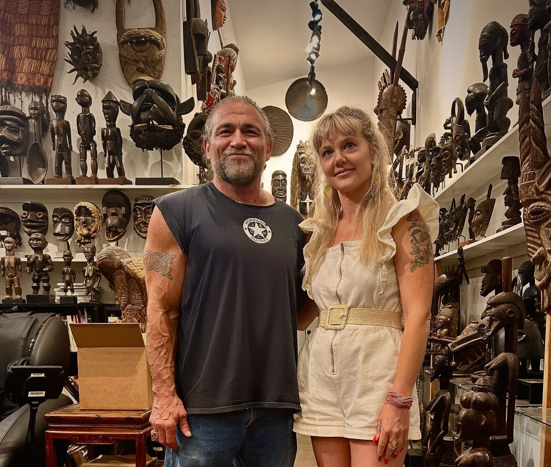 Two people standing in a shop filled with hanging masks and carvings, smiling at the camera.