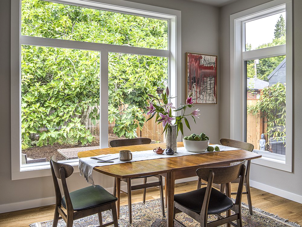 Bright dining room with large windows, wooden table, chairs, and a vase of flowers overlooking greenery
