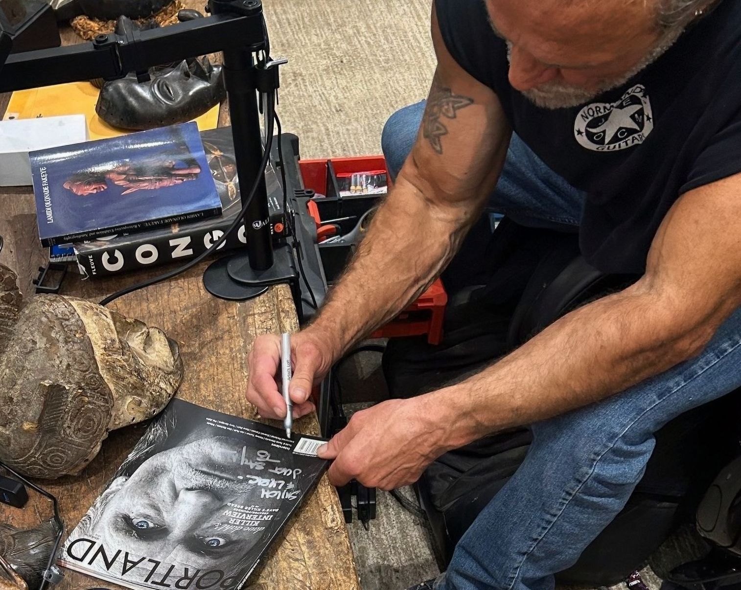 Man signing a black-and-white poster at a table, with a rock and blue case nearby