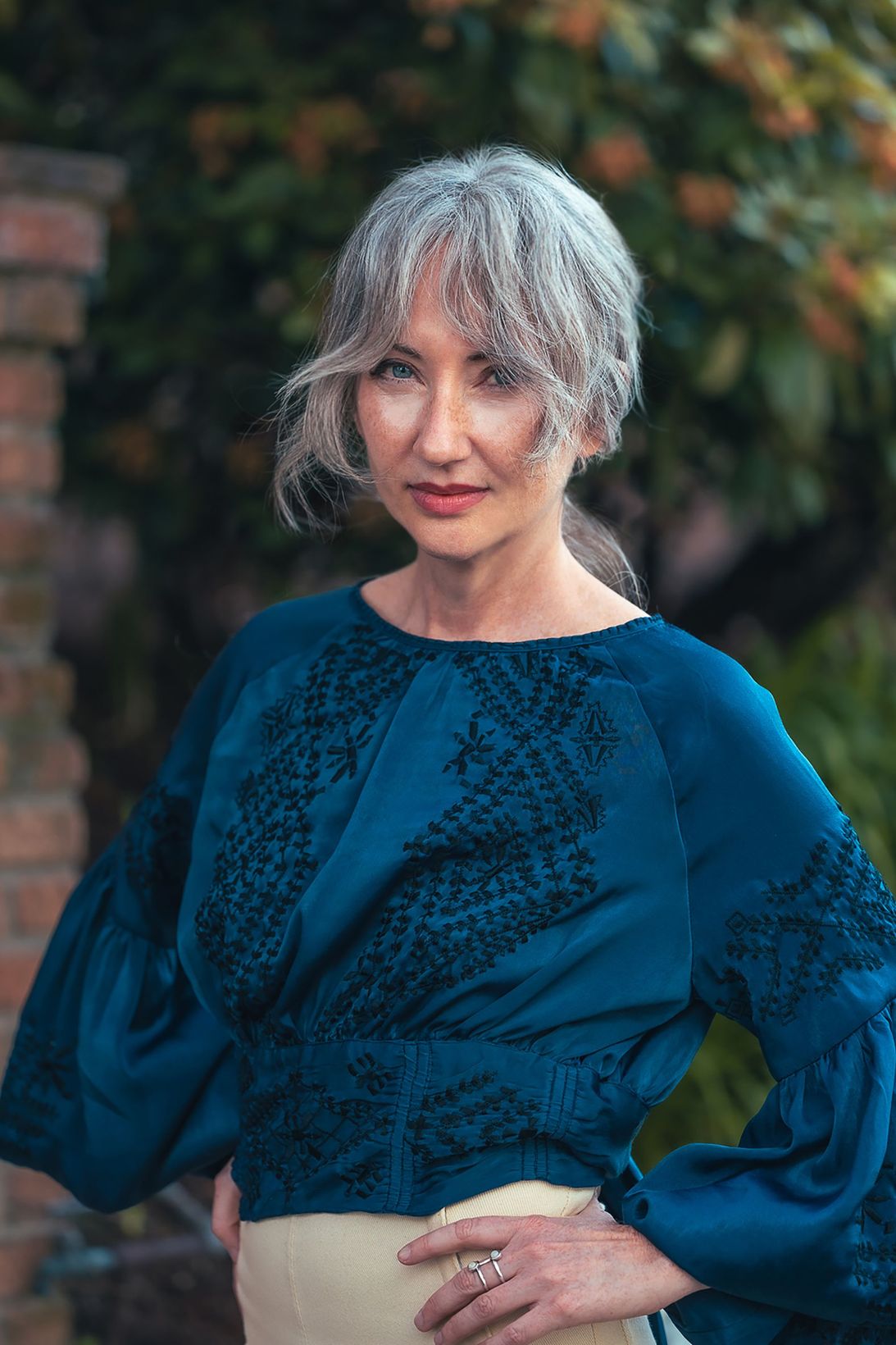 Person with silver hair wearing a teal blouse and beige skirt, standing outdoors with greenery in the background.