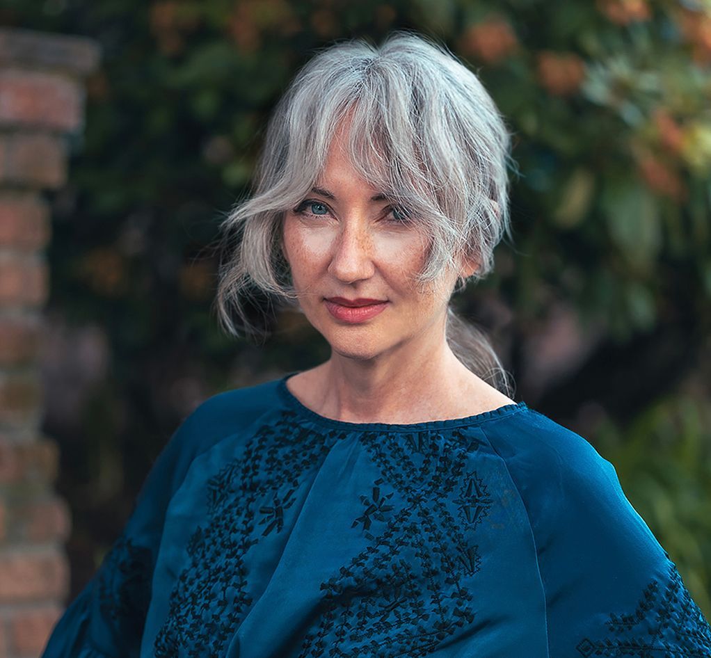 Portrait of a woman in a blue top with silver hair, standing outdoors with blurred greenery behind her