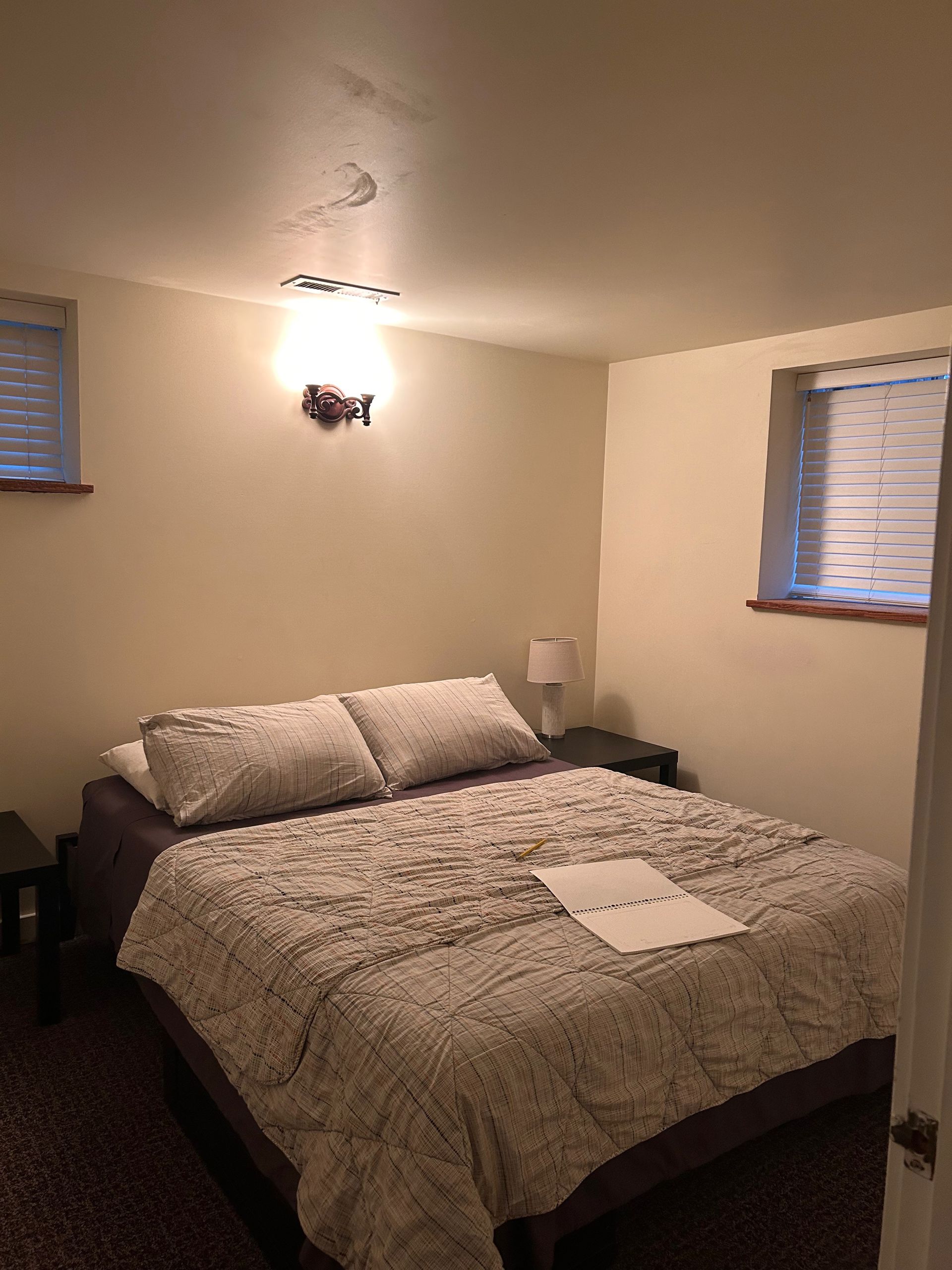 Small bedroom with a bed, patterned bedding, wall sconce, and two windows with blinds.