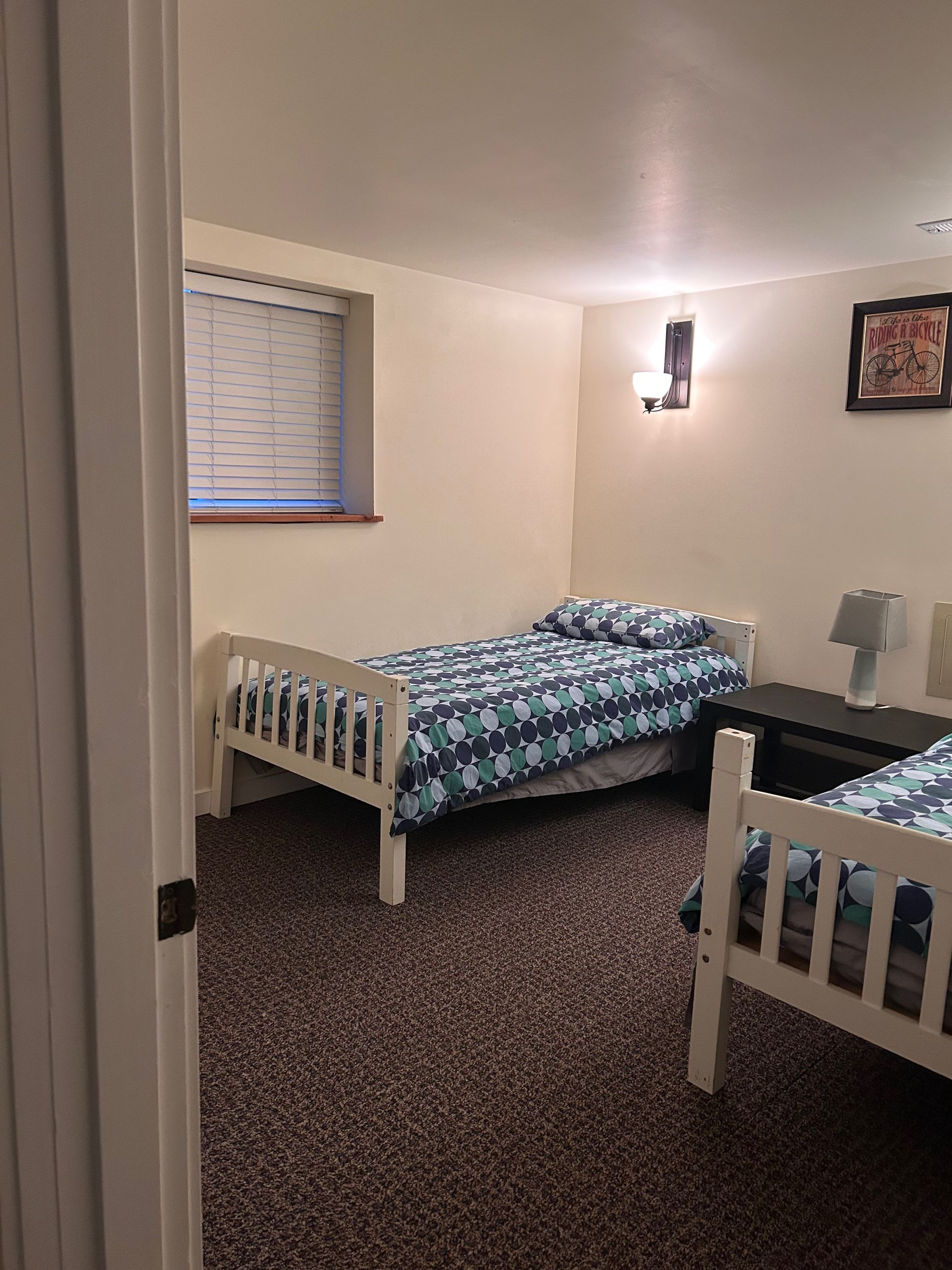 Small bedroom with two white twin beds, beige walls, and dark patterned carpet.