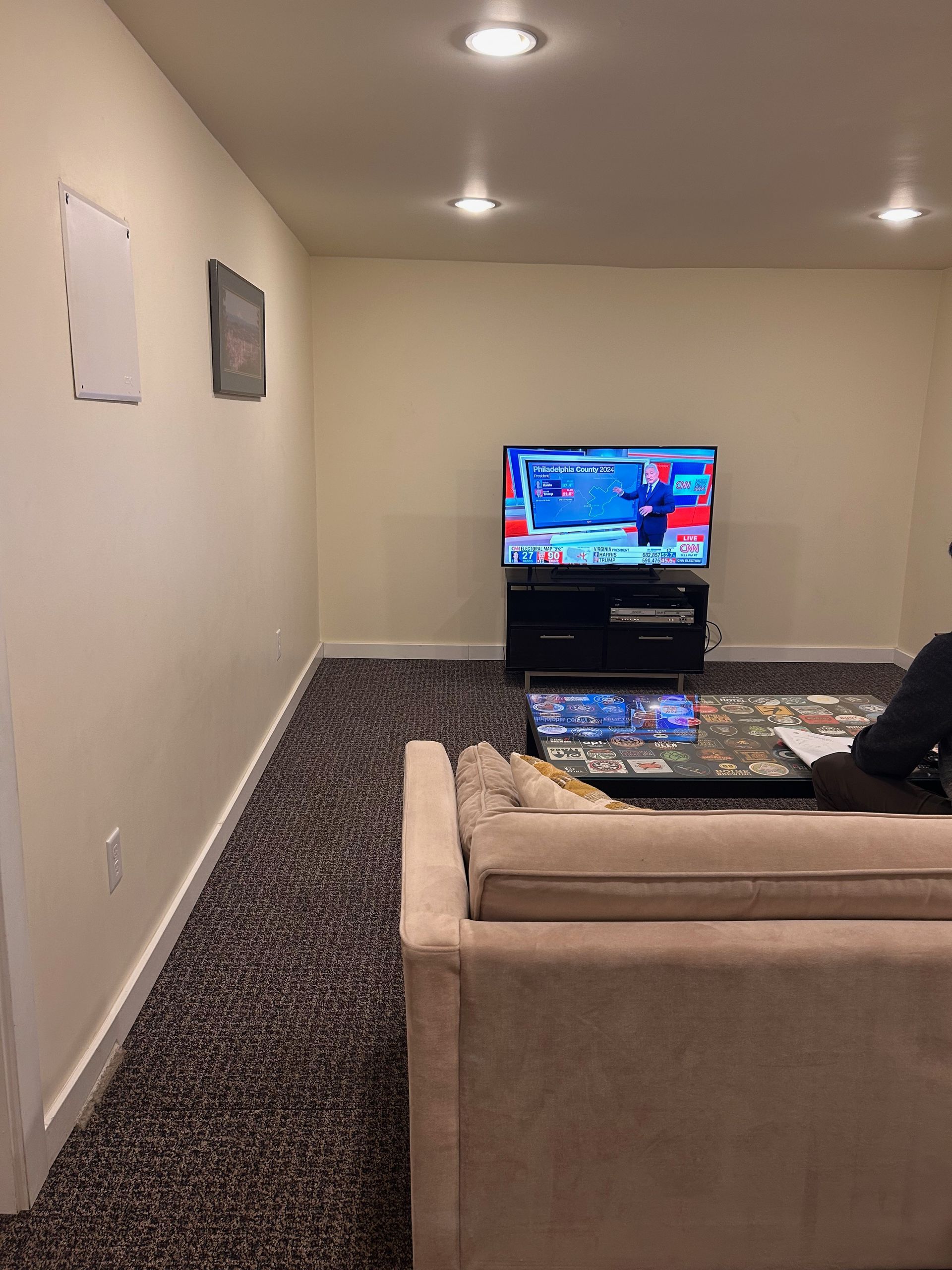 Living room with beige couch facing a TV on a stand, dark carpet, and recessed ceiling lights