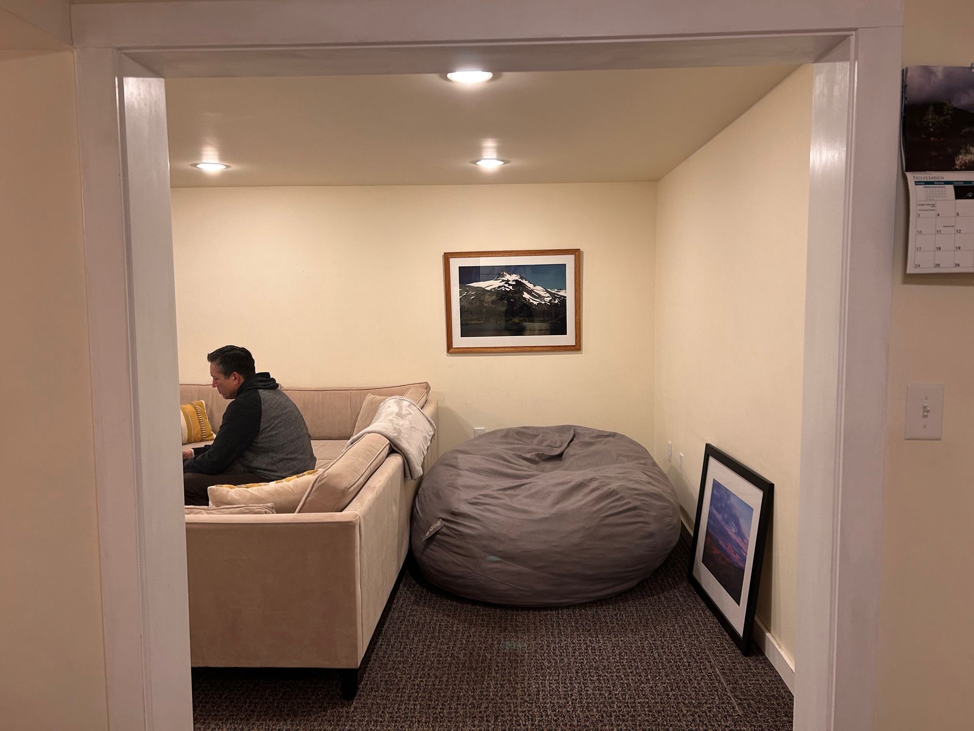 Cozy living room with beige couch, gray bean bag, framed wall art, and a person sitting on the sofa.