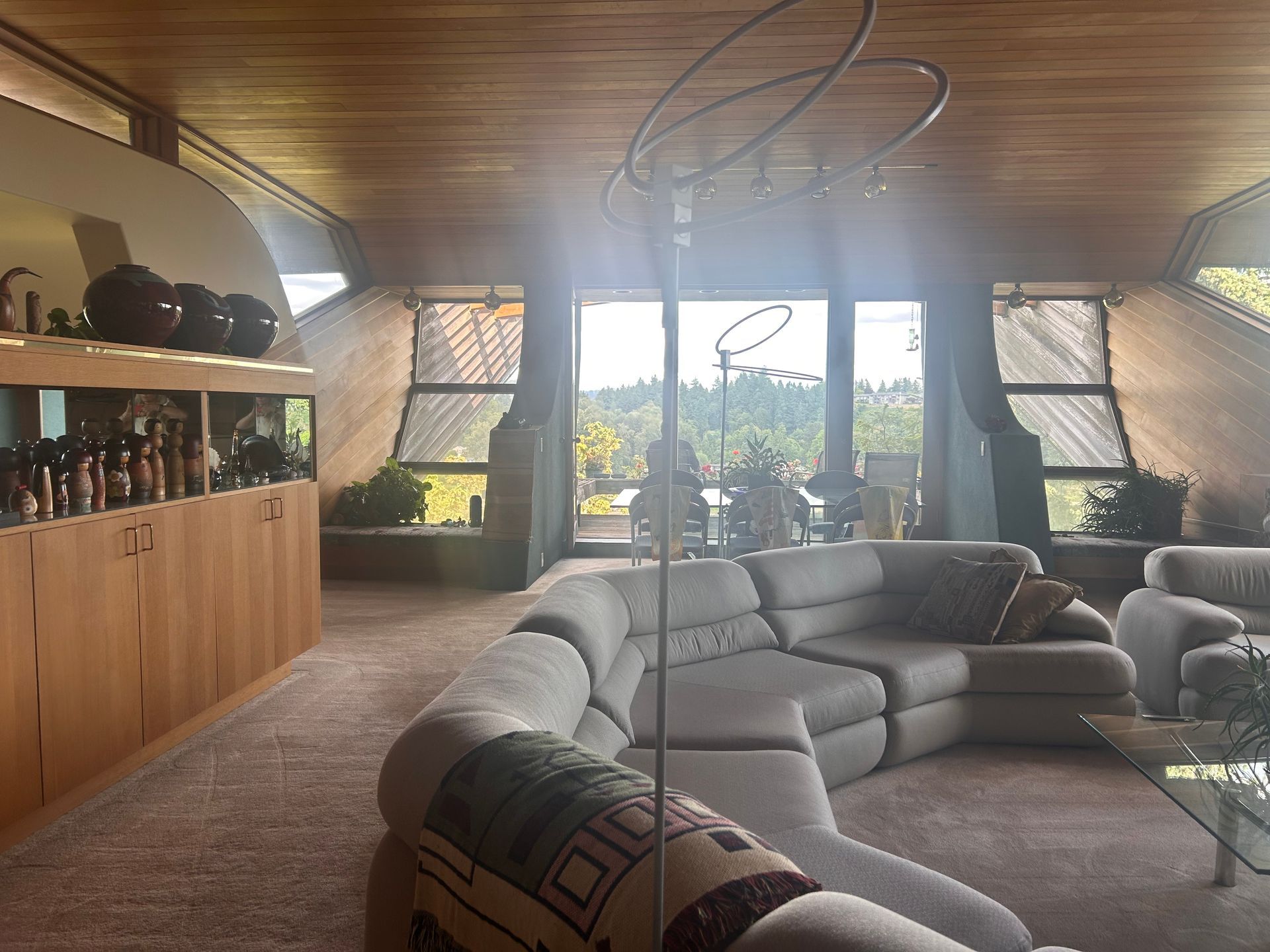 Open-air living room with sofas, wooden ceiling, and garden view through sliding doors.