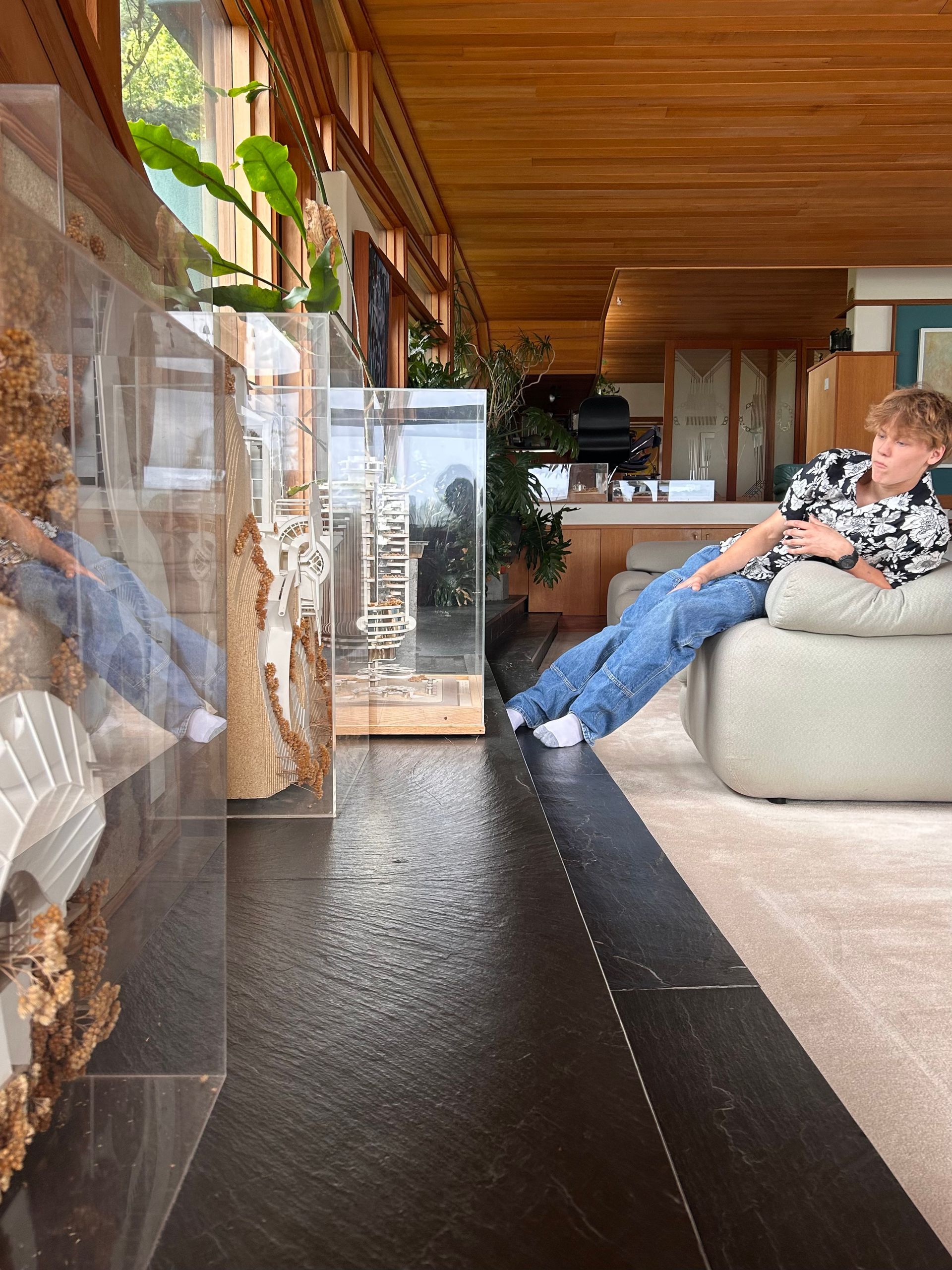 Modern living room with a person reclining on a white sofa beside a glass wall and wooden ceiling.